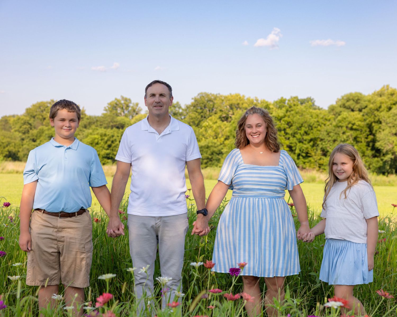 Family of four holding hands in a wildflower meadow on a sunny day, smiling outdoors.