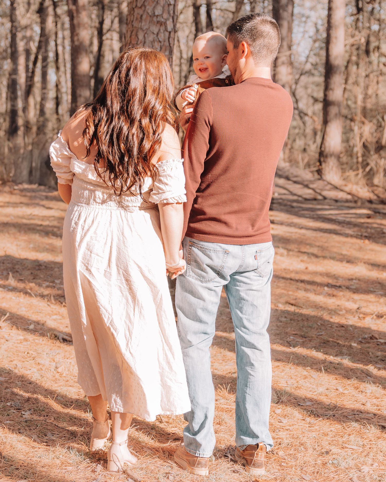 A couple in casual attire holds their baby while walking through a wooded area.