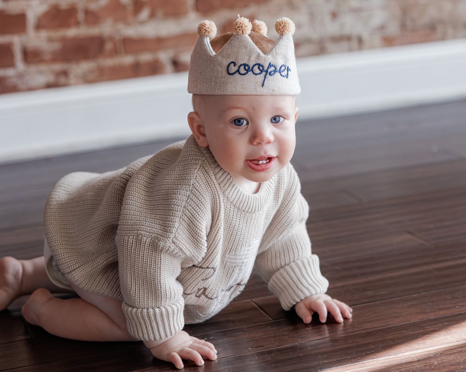 Baby wearing a personalized knit crown hat with the name Cooper, crawling on a hardwood floor in a cream sweater.