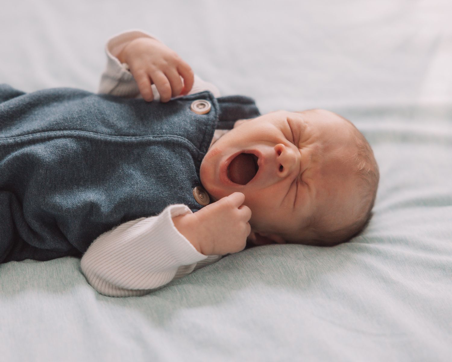 Yawning newborn baby in blue romper lying on a white blanket with tiny fists raised.