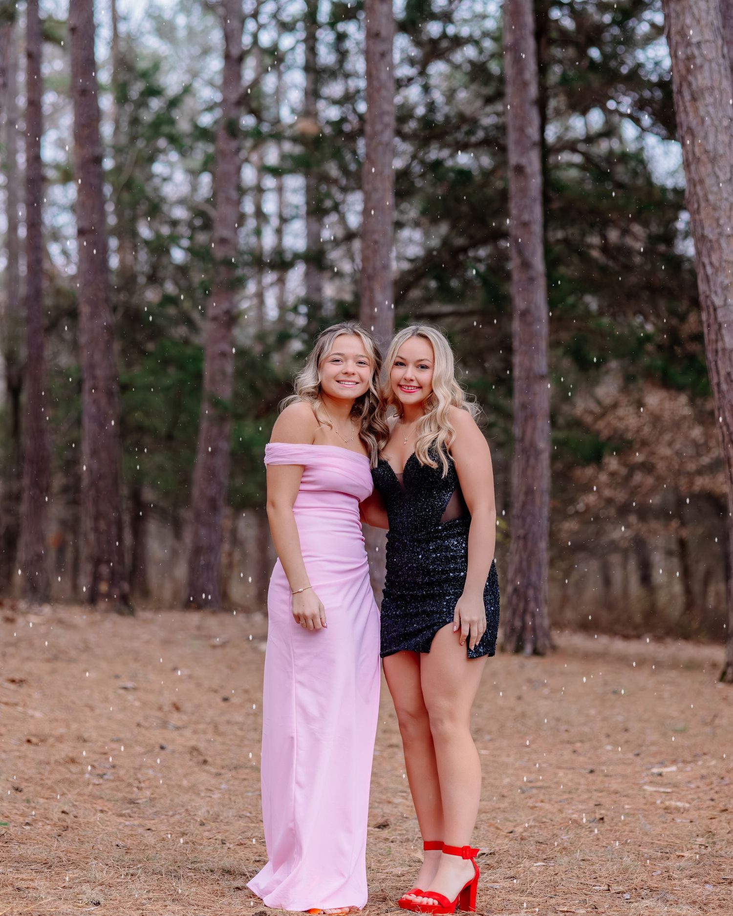 Two people in formal attire pose together in a wooded setting, one in a pink gown and one in a black dress.