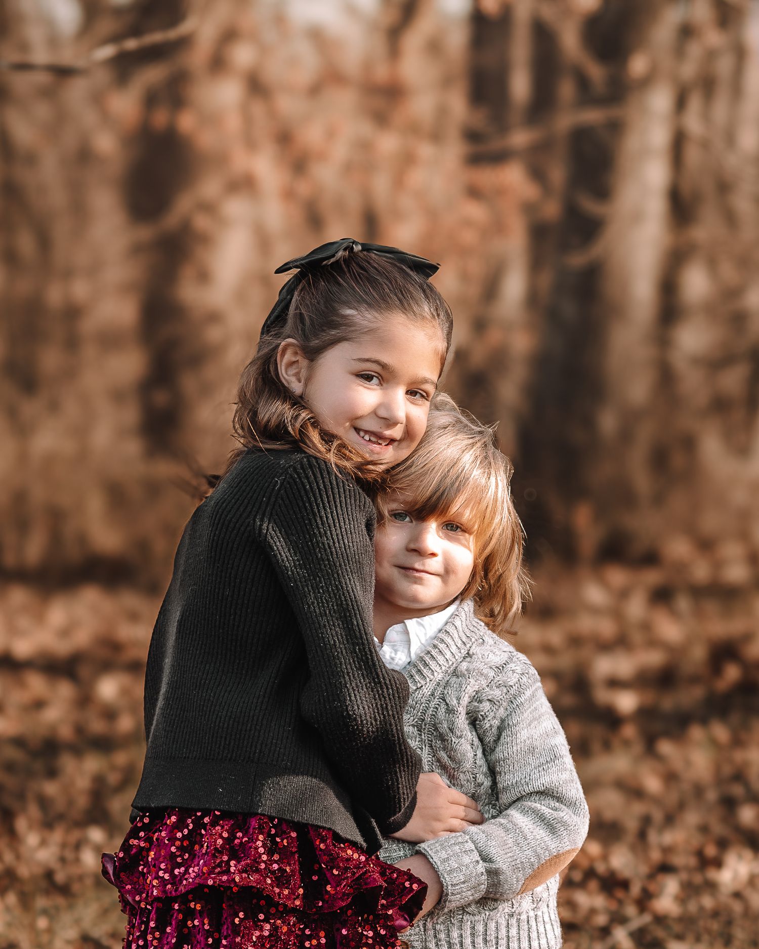Two people share a warm embrace while sitting among fallen autumn leaves in a forest setting.