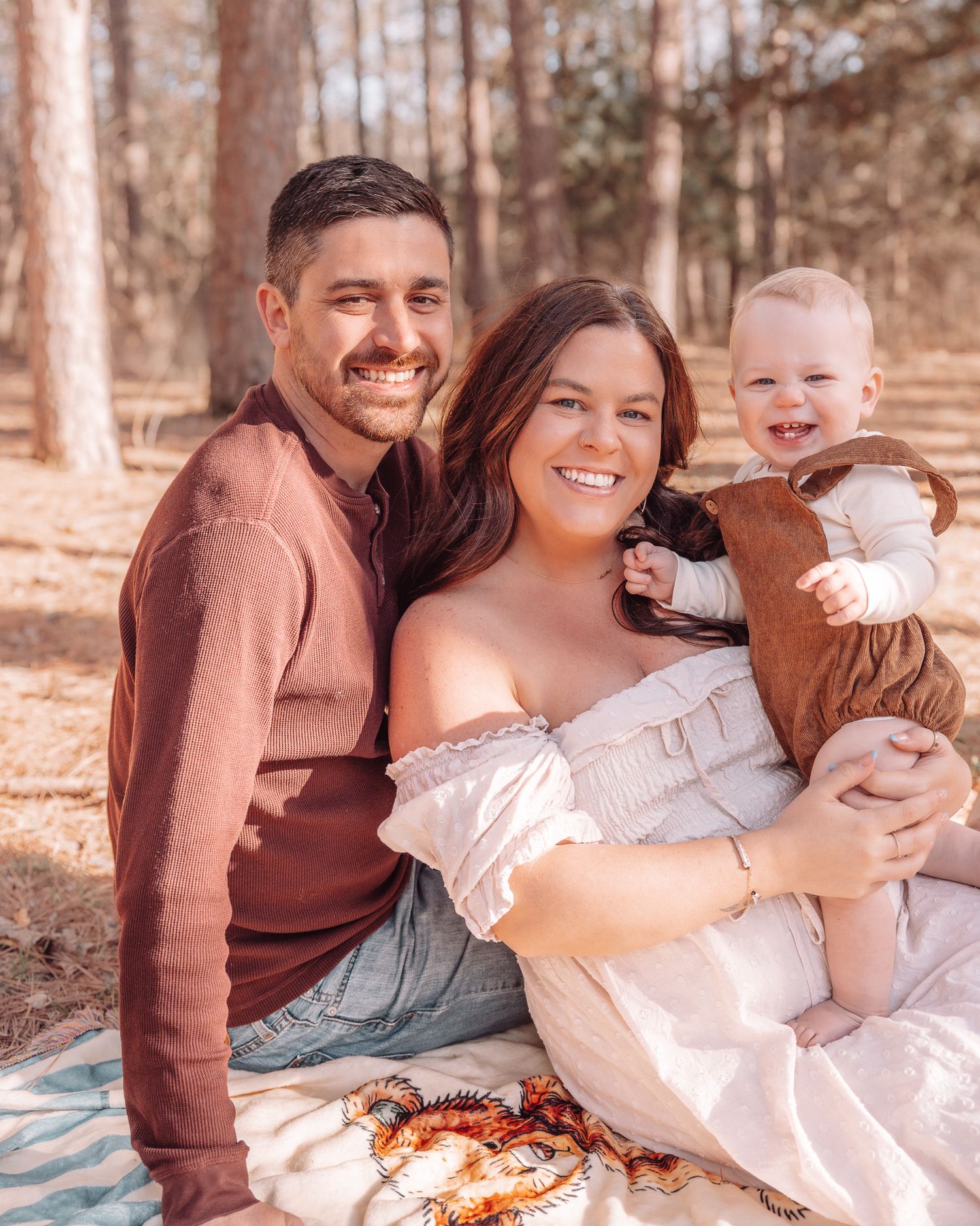 A happy young family with a smiling couple and baby sitting outdoors among pine trees on a colorful blanket.