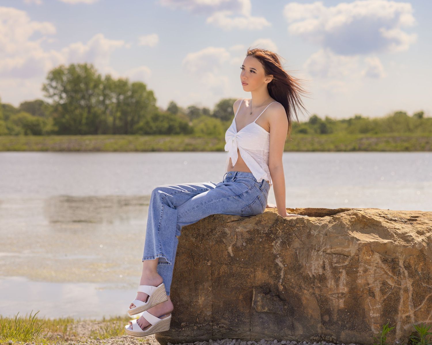 Young woman in jeans and white top sitting on a rock by a lake on a sunny day.