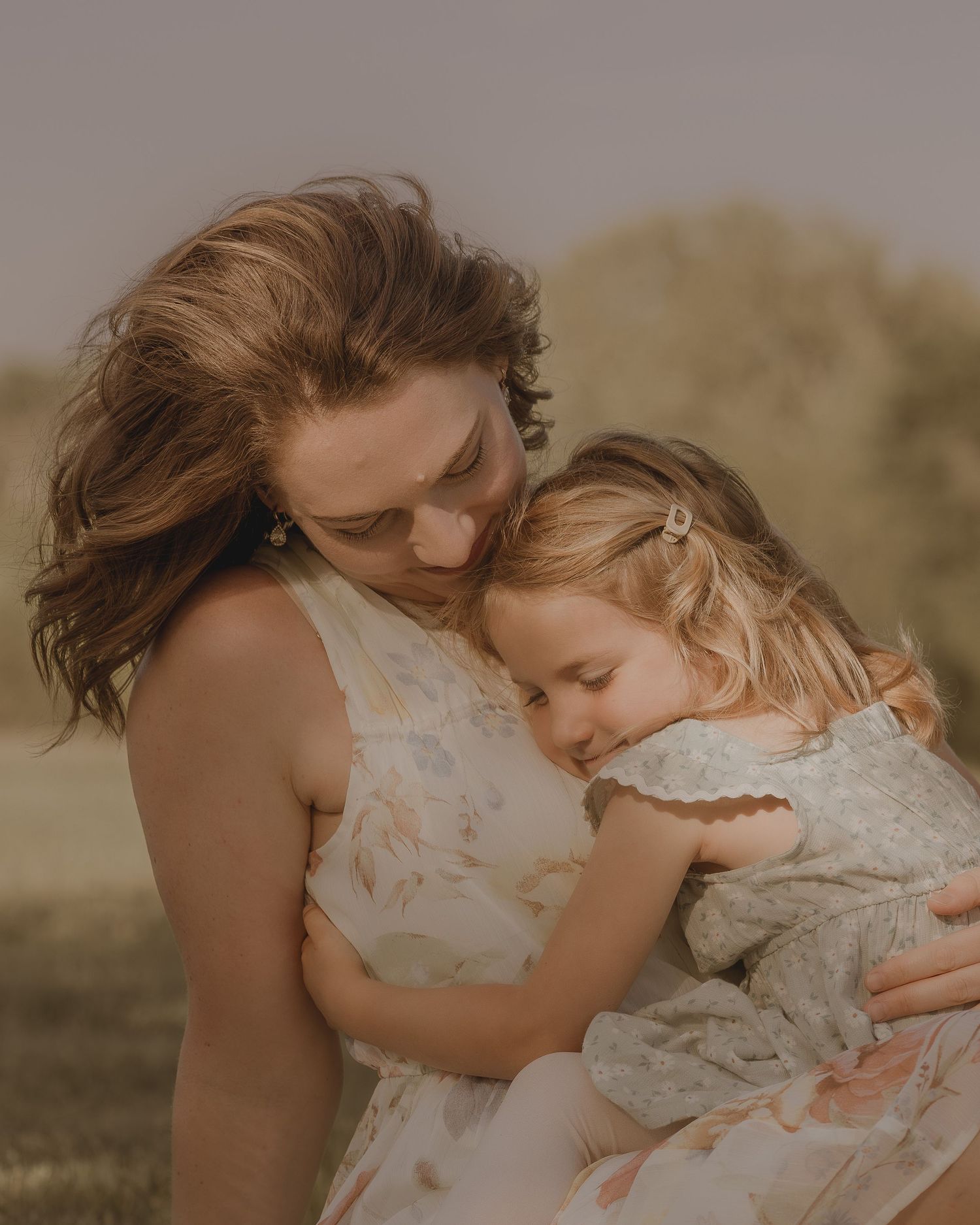 A tender moment as a mother gently embraces her young daughter outdoors in a soft golden light setting.
