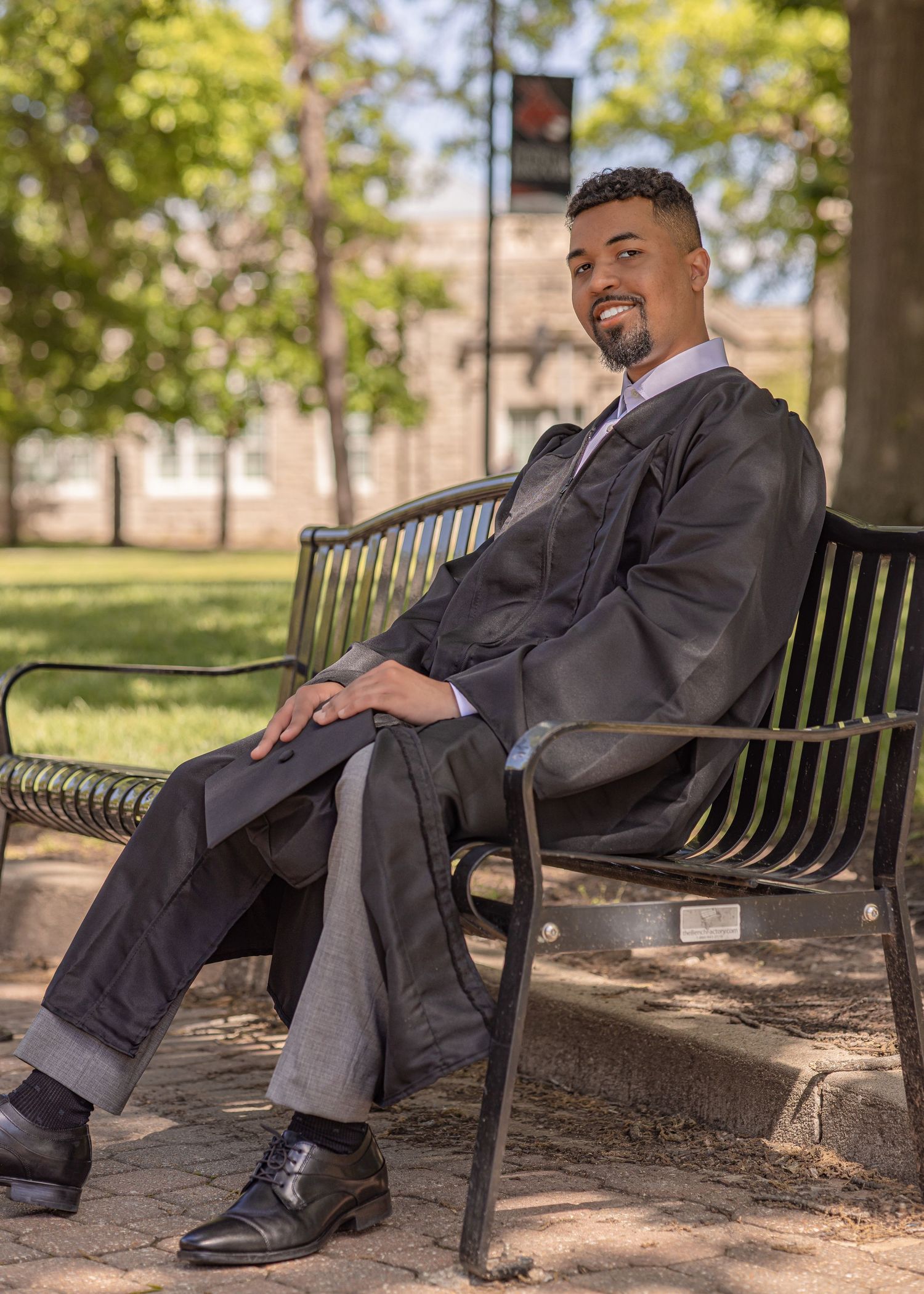 Man in graduation gown sitting relaxed on a park bench outdoors, smiling confidently in formal attire.