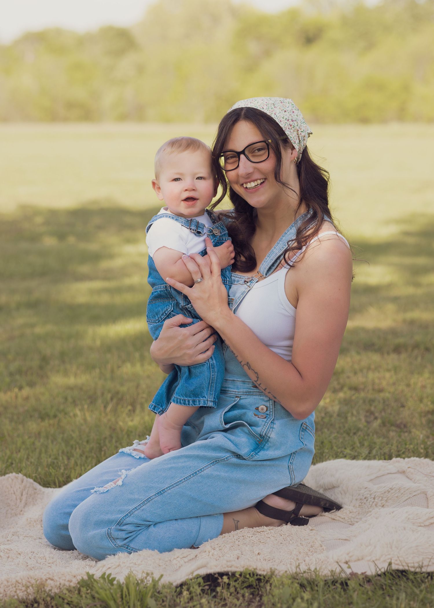Smiling woman in denim overalls and glasses holds baby outdoors on blanket in sunny green park.