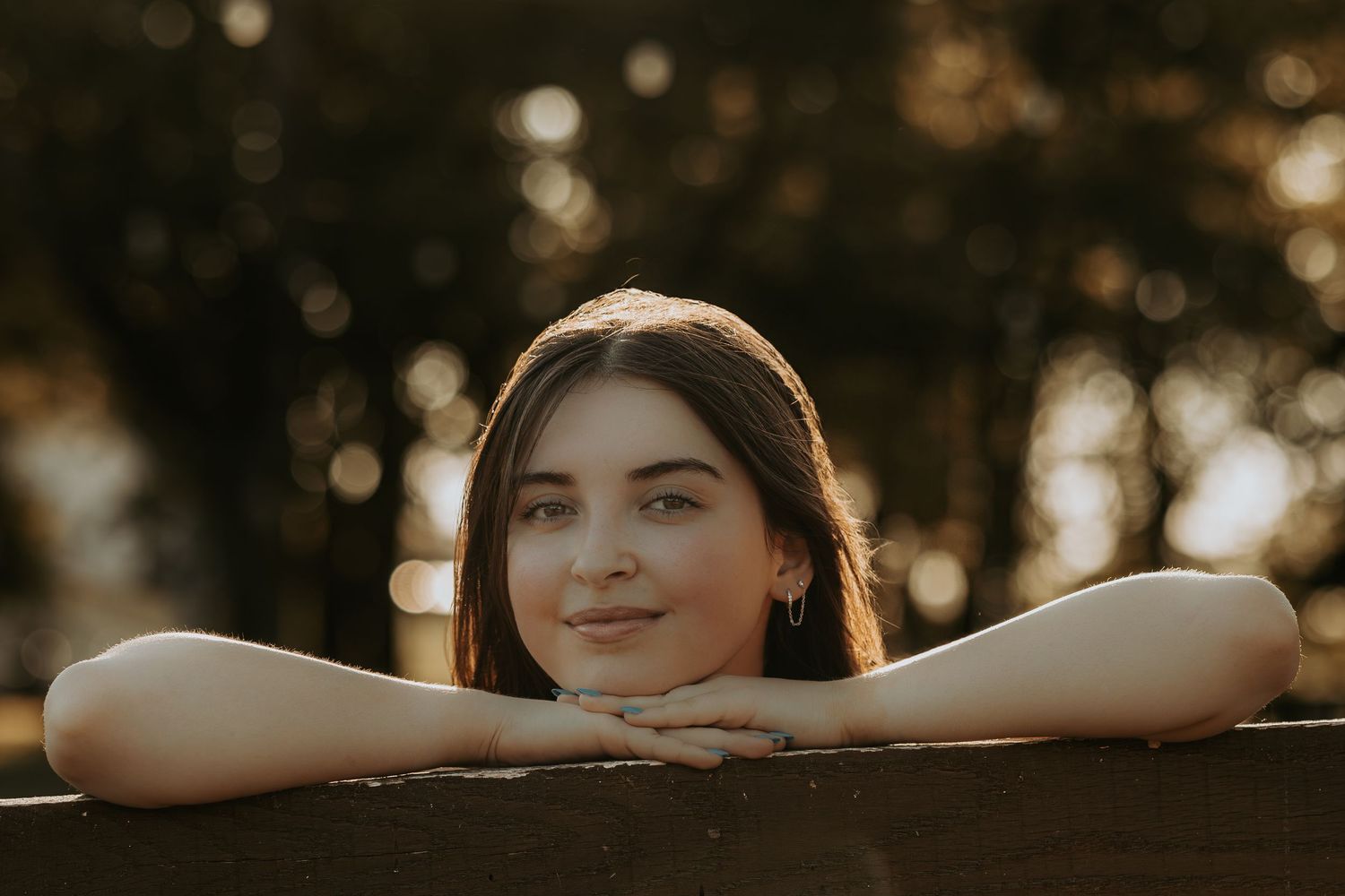 A backlit portrait of a young woman resting her chin on a wooden railing during golden hour.