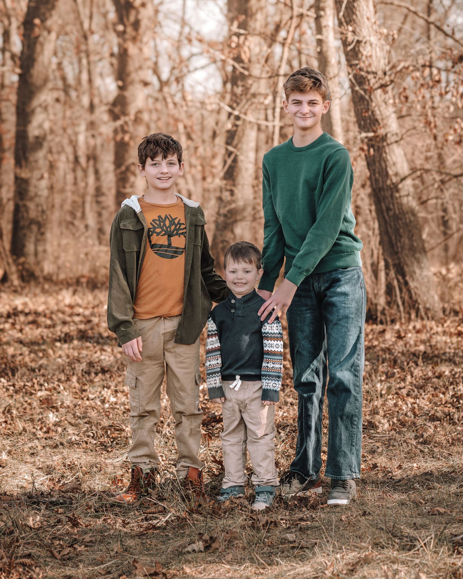 A family in coordinated fall outfits walks through a wooded area with fallen autumn leaves on the ground.