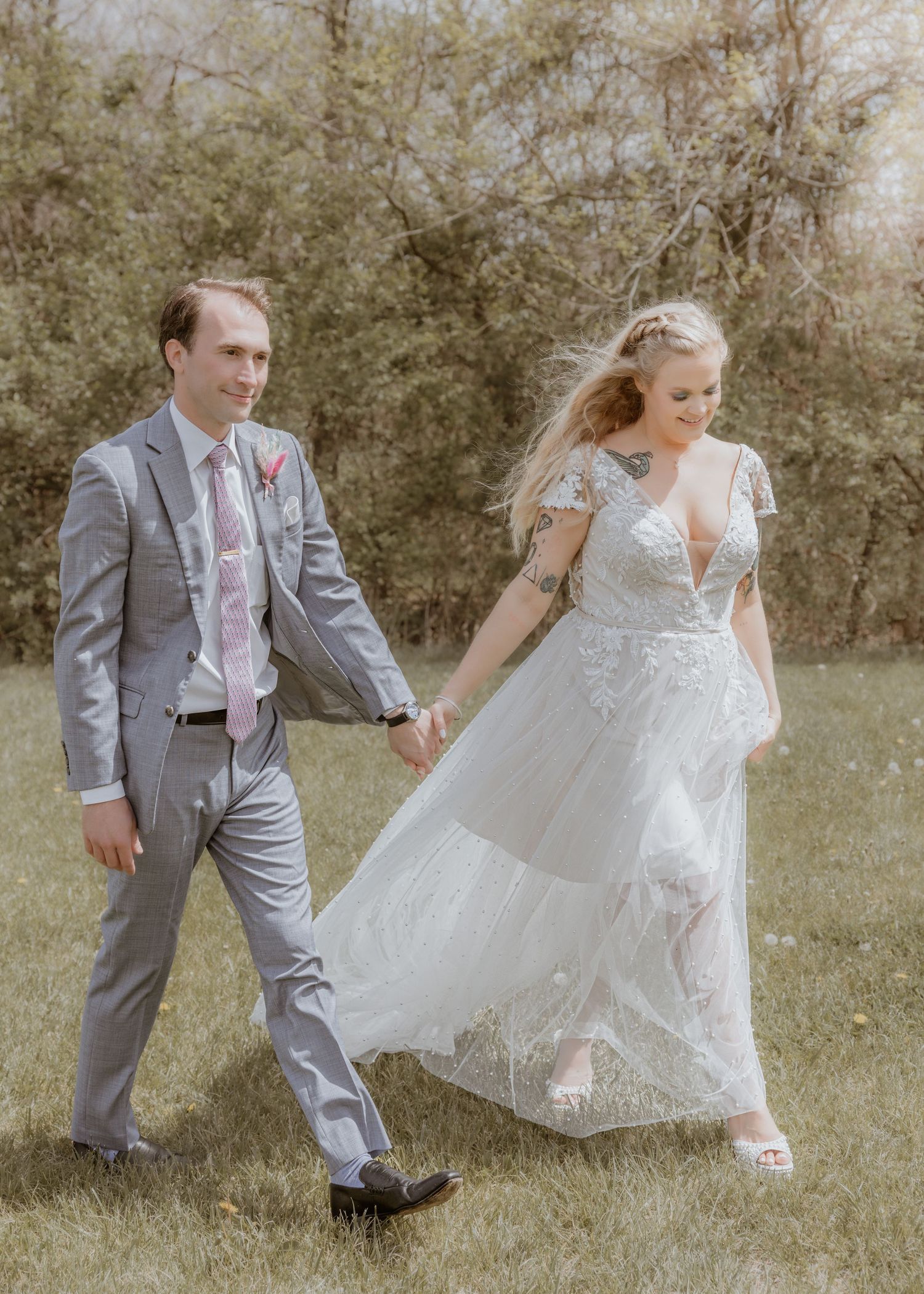 Groom in gray suit holds hands with bride in lace gown walking across green lawn.