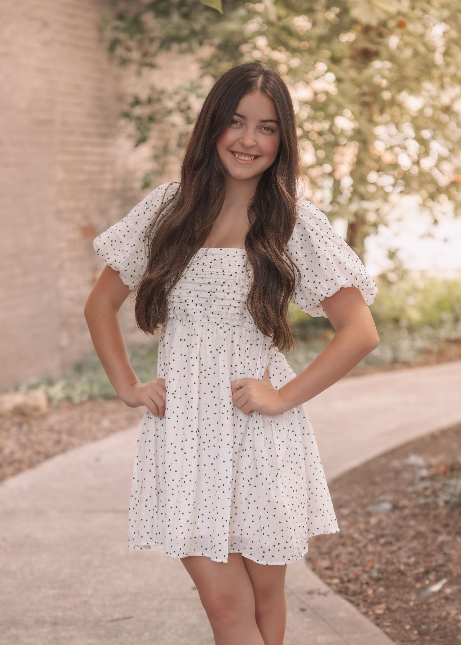 Young woman smiling in a white floral mini dress with puff sleeves, posing outdoors near a brick wall.
