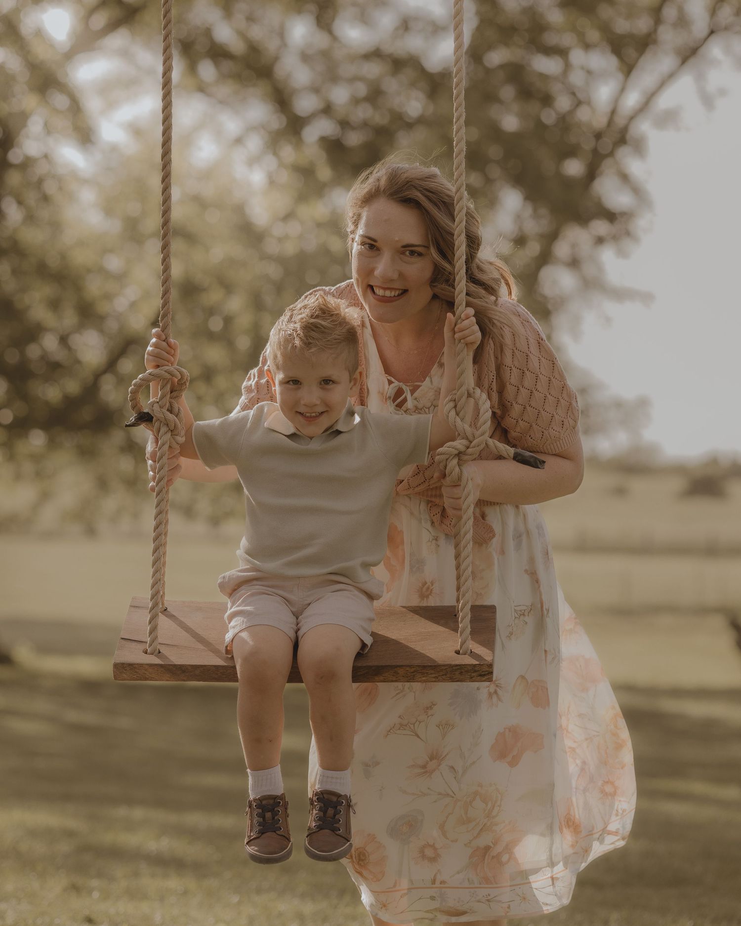 A smiling mother in a floral dress sits with her young son on a wooden rope swing outdoors.