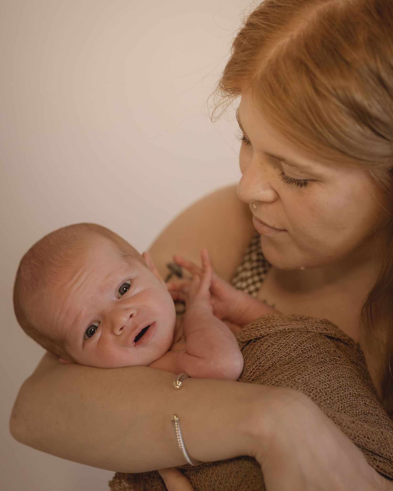 A red-haired mother tenderly holds her newborn baby wrapped in a brown knit blanket against a neutral background.