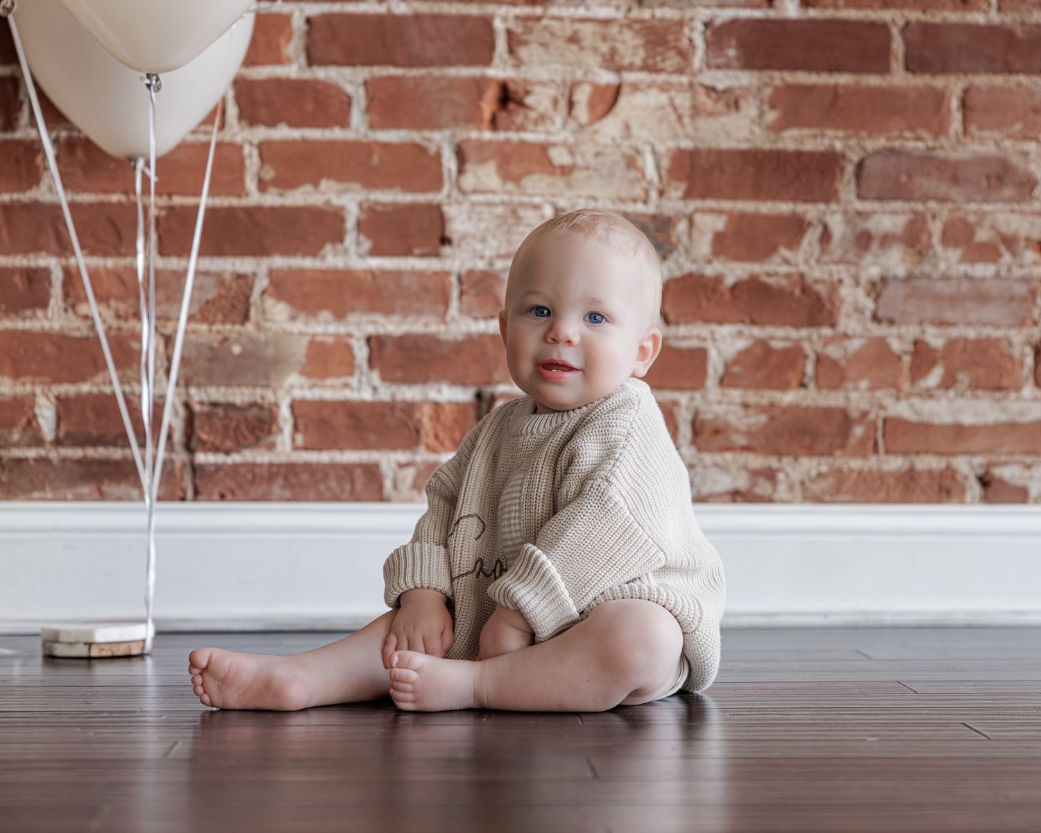 Adorable baby sitting on hardwood floor in cream knit romper with white balloons and brick wall backdrop.