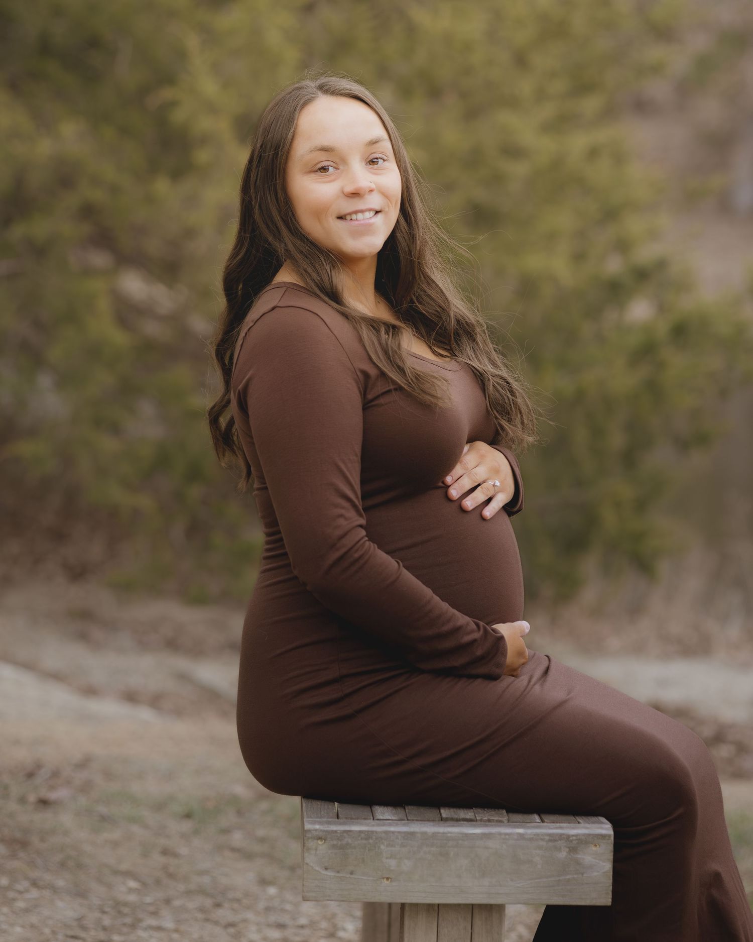 Pregnant woman in brown long-sleeve dress sitting on bench outdoors, smiling, cradling baby bump during maternity photo session.