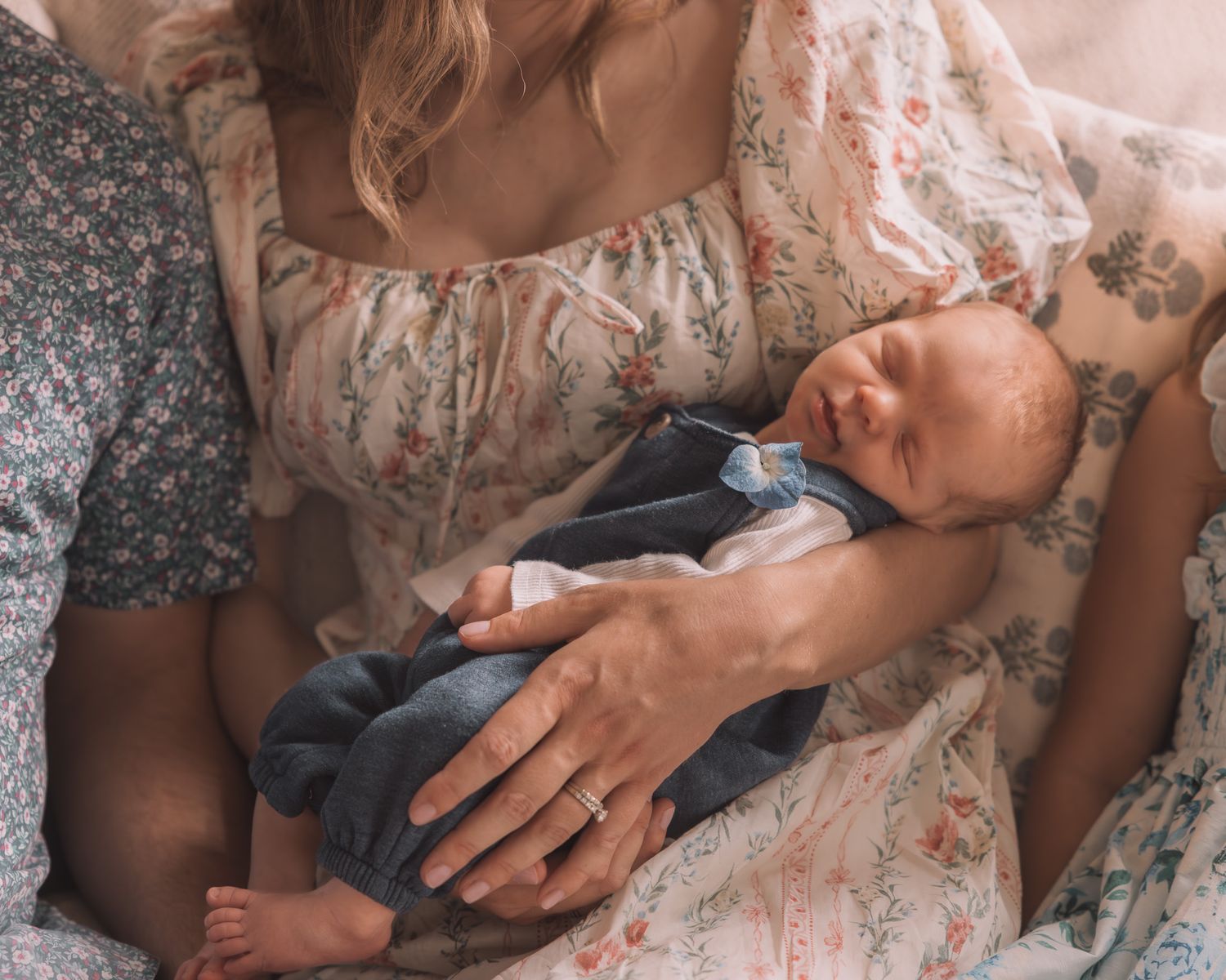 Mother cradling sleeping newborn baby dressed in blue outfit while sitting on a floral bed.