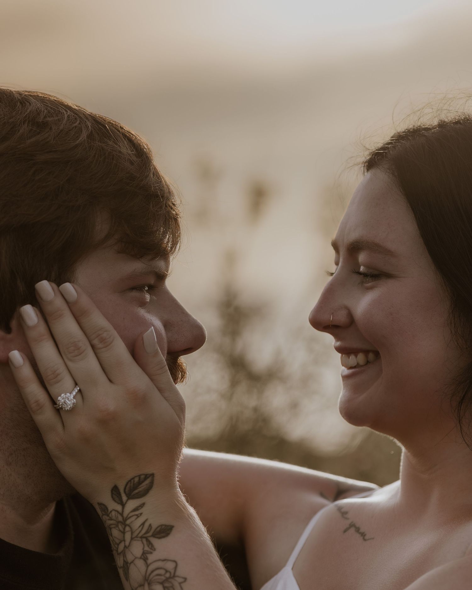 Couple sharing a romantic moment, woman with floral tattoo and engagement ring gently touching mans face, smiling softly.