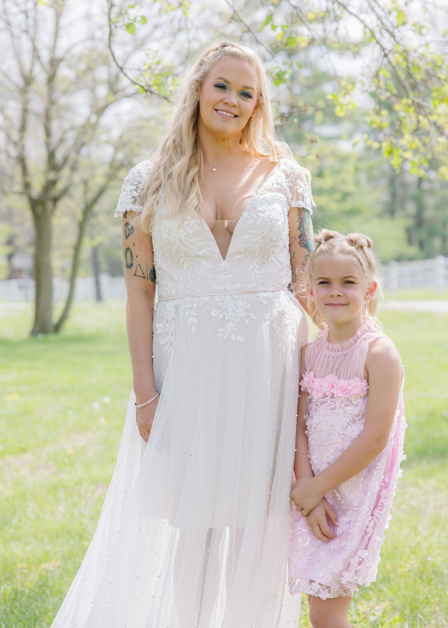 Bride in white lace gown poses with young girl in pink dress outdoors among spring trees.