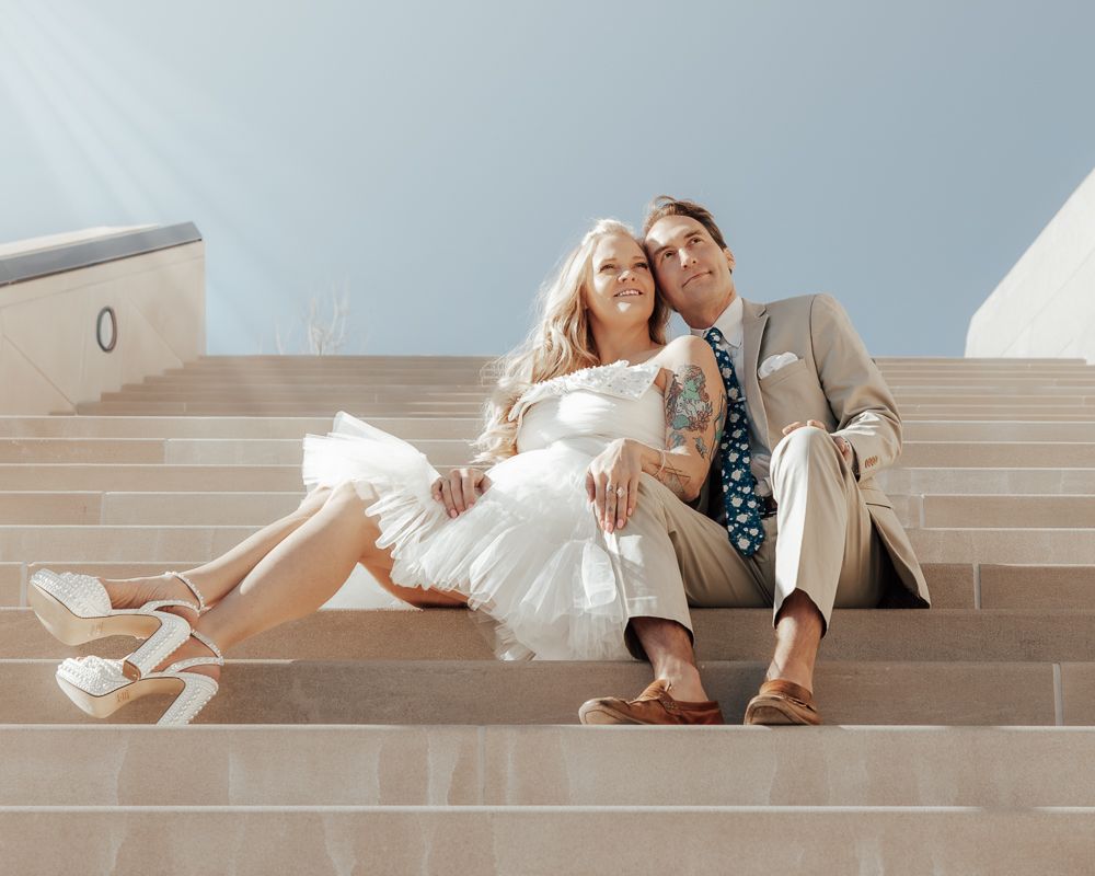 A romantic couple in formal attire poses on light-colored steps during a wedding photoshoot.