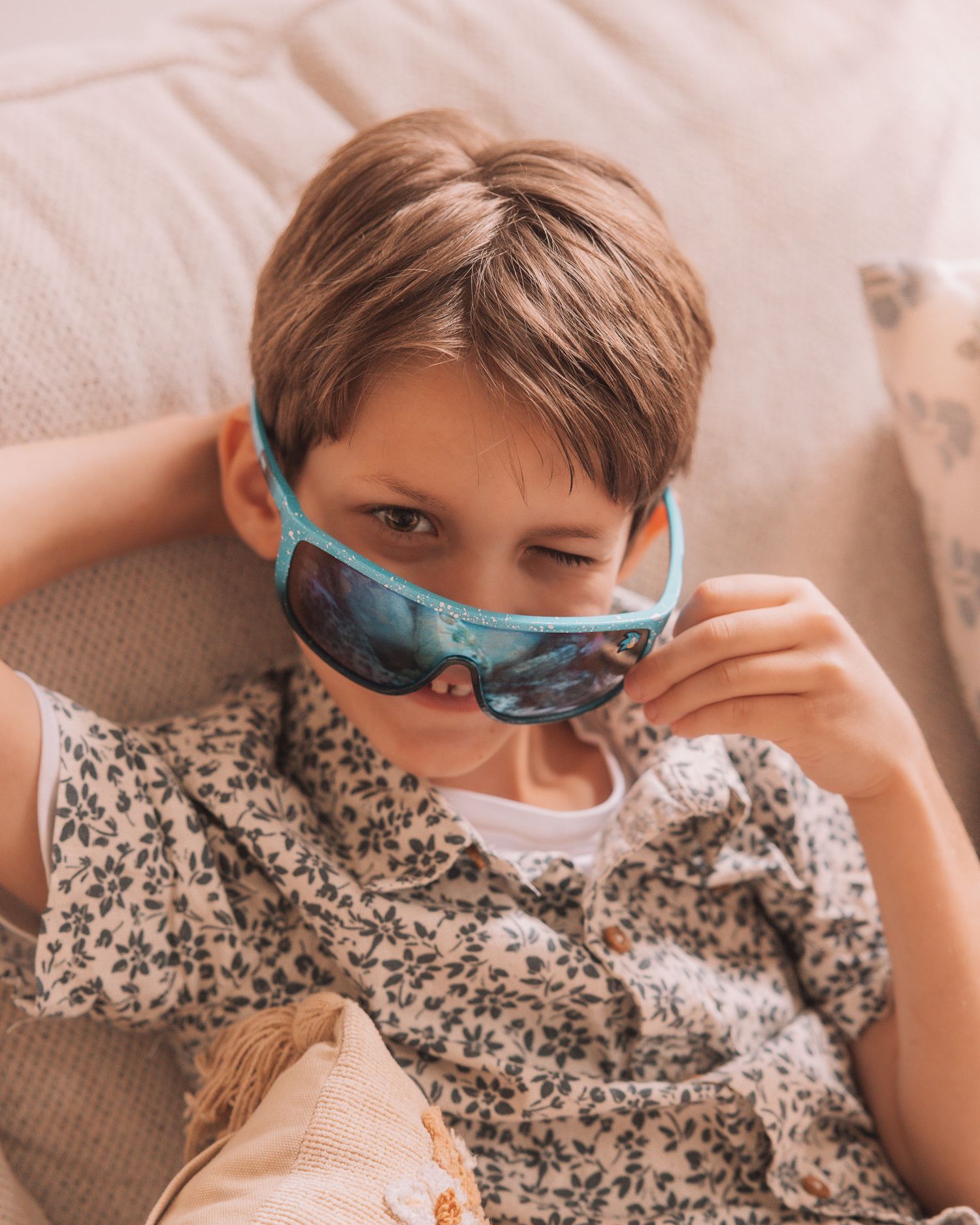 Young boy wearing teal Frozen sunglasses lounging on a beige couch in a floral shirt.