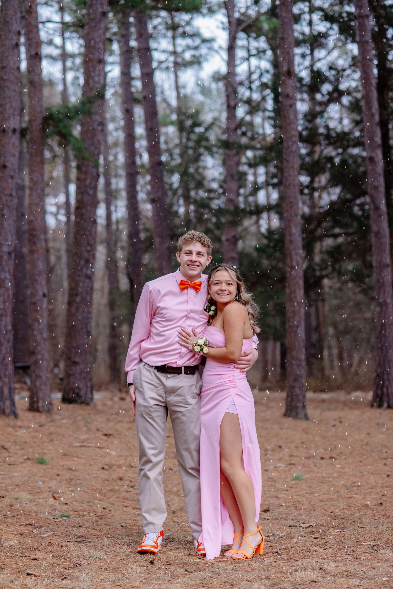 A couple in pink attire shares romantic moments in a pine forest setting during a photo session.