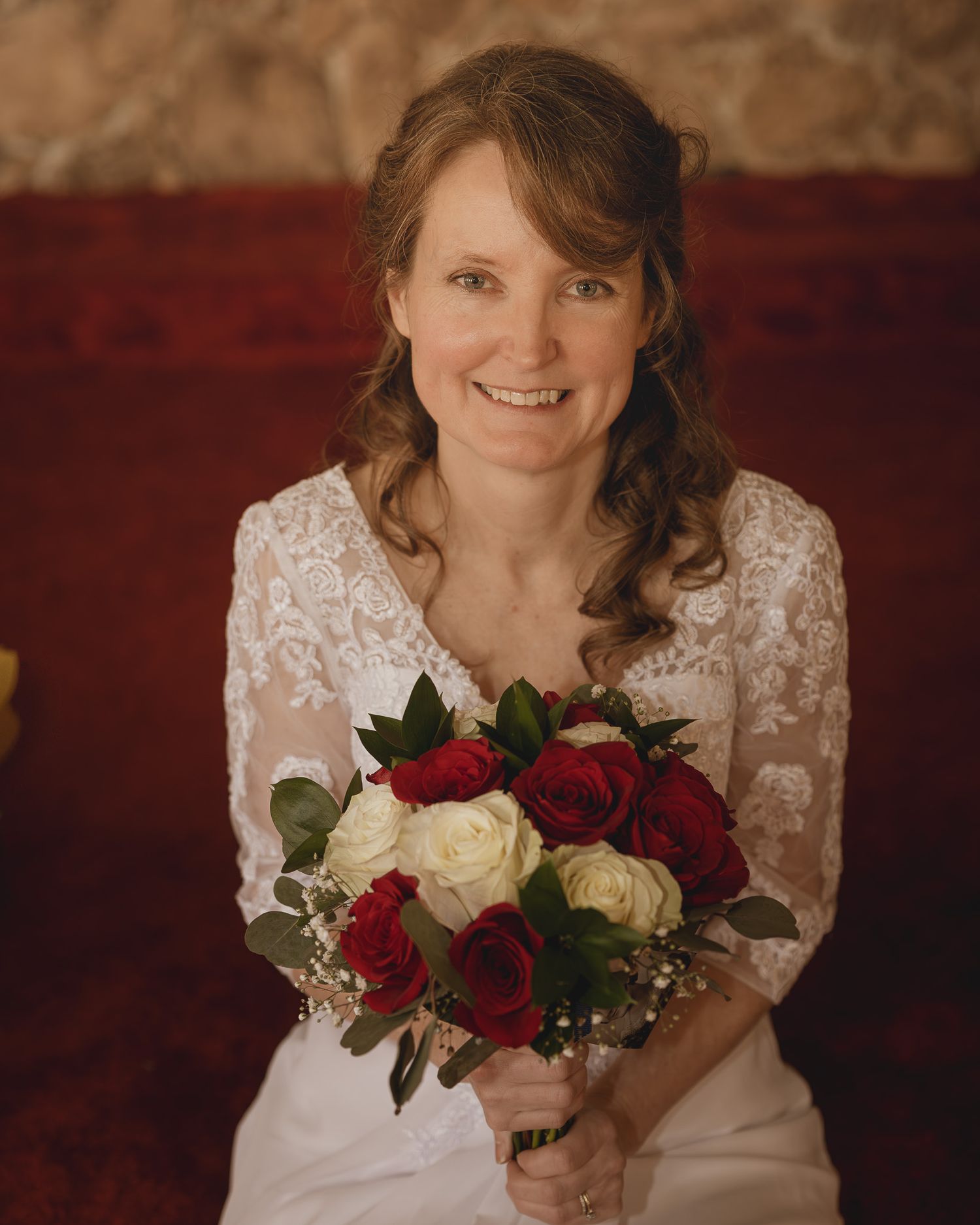 A bride in a white lace dress holds a bouquet of red and white roses against a dark red background.