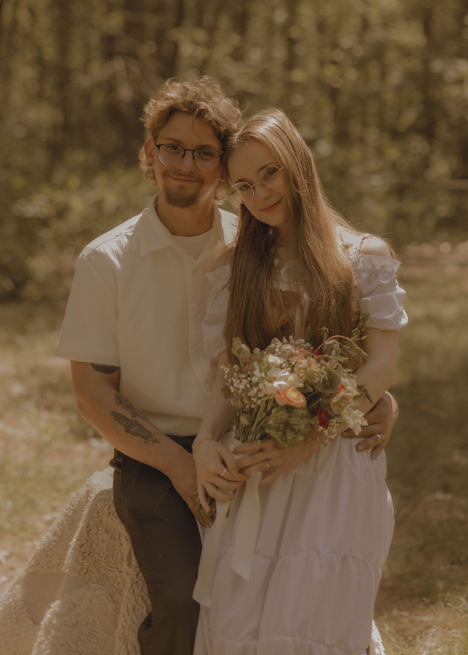 A couple in wedding attire poses in a forest, the bride holding a floral bouquet in a white dress.
