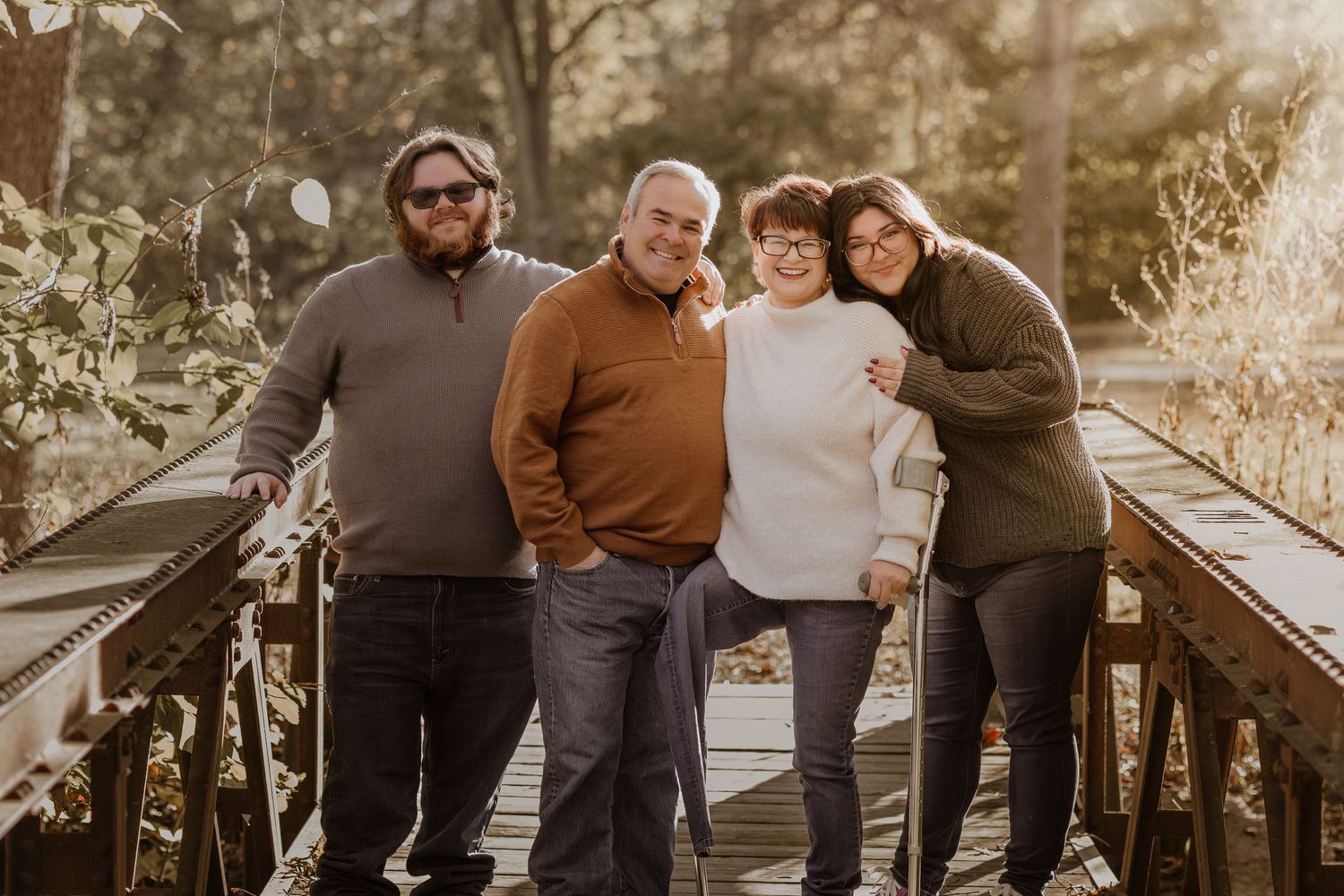Family of four smiling together on a wooden bridge surrounded by autumn foliage in warm natural light.