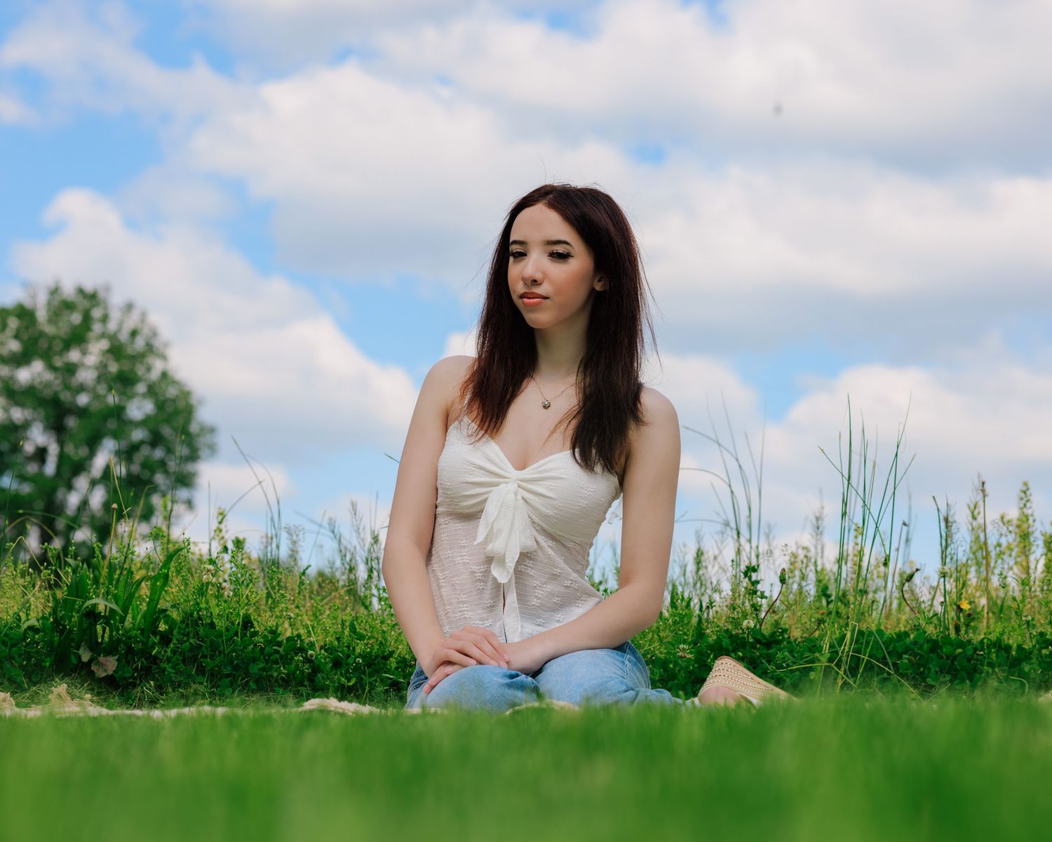 Young woman in white top and jeans sitting on green grass field under a blue cloudy sky.