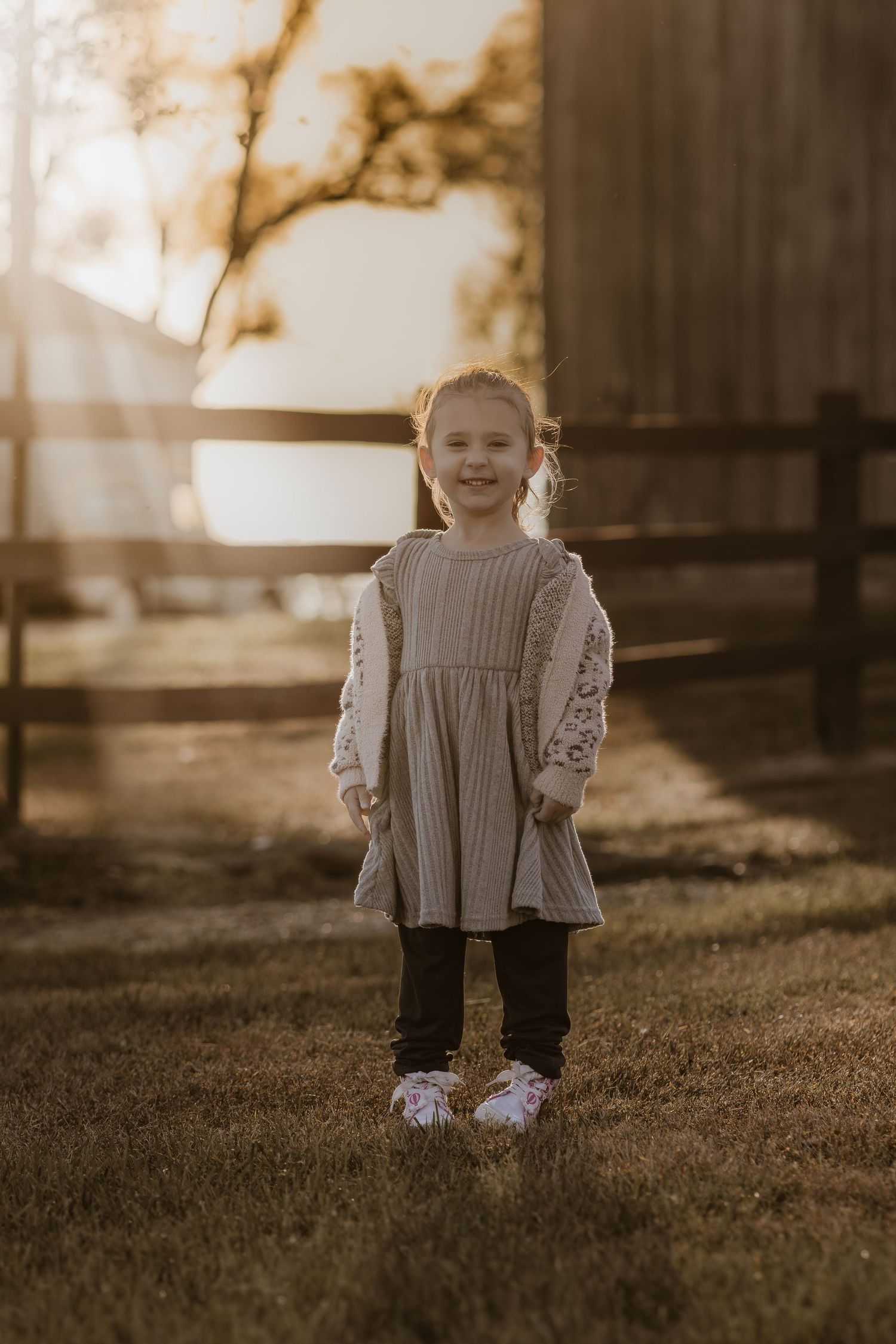 A toddler in a gray sweater dress stands on grass during sunset.