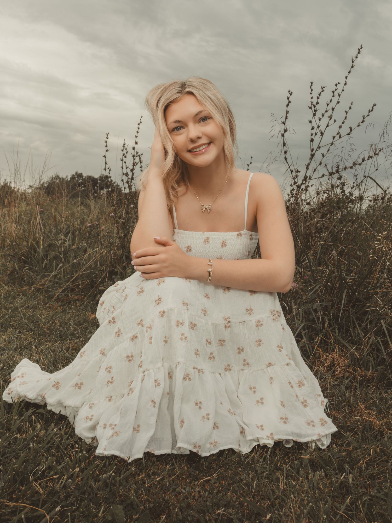 Blonde woman in white floral sundress sitting in a field with overcast sky.