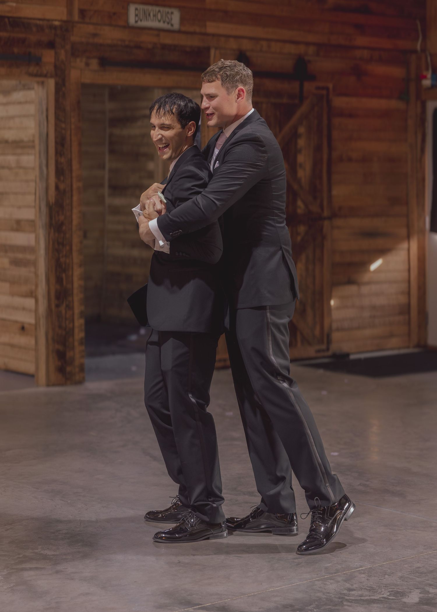 Two men in dark suits laughing and embracing on a dance floor inside a rustic wooden barn venue.