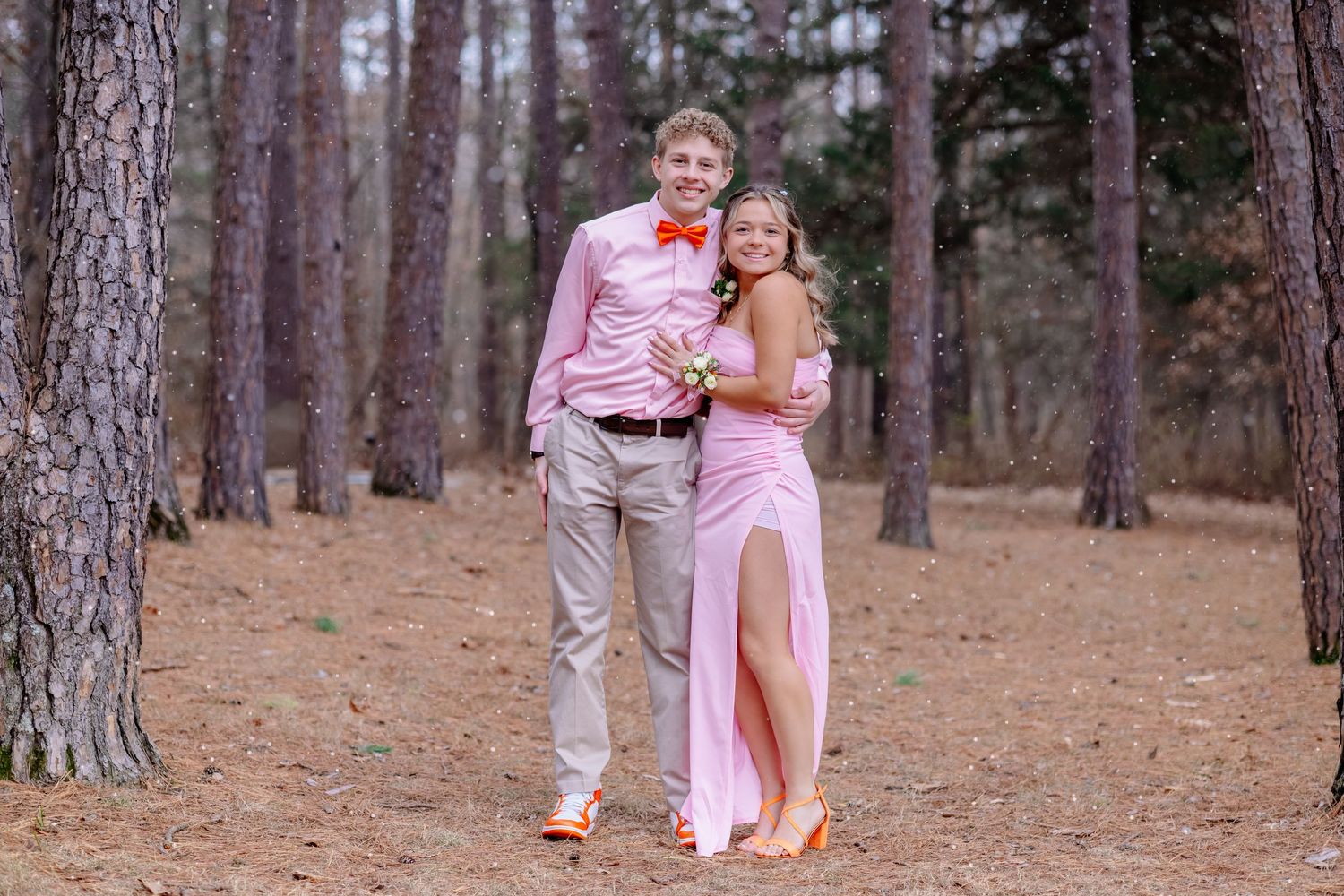 A couple in coordinating pink outfits pose together in a pine forest with brown needles on the ground.