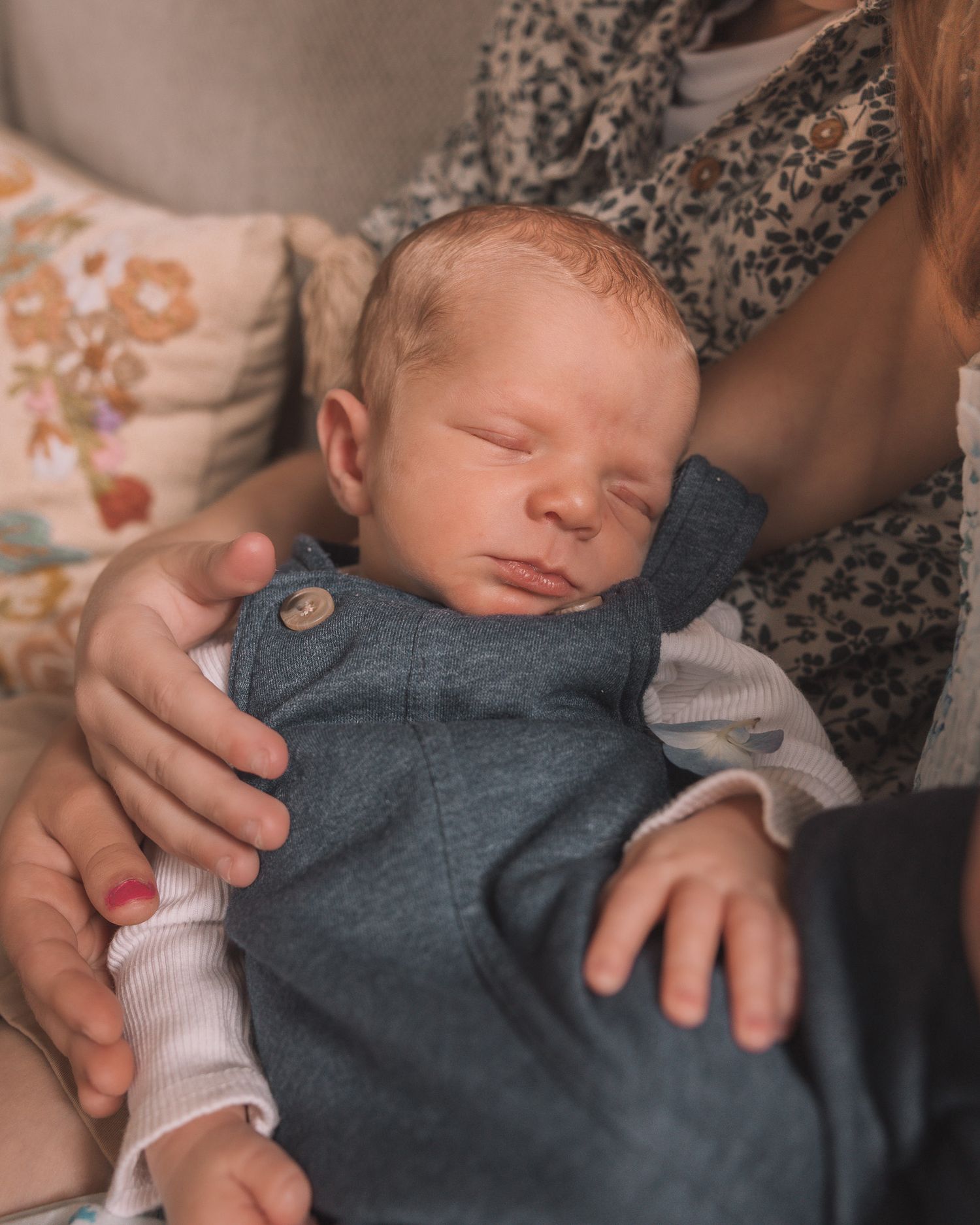 Sleeping newborn baby with red hair in gray overalls being held by a mother in a floral dress.