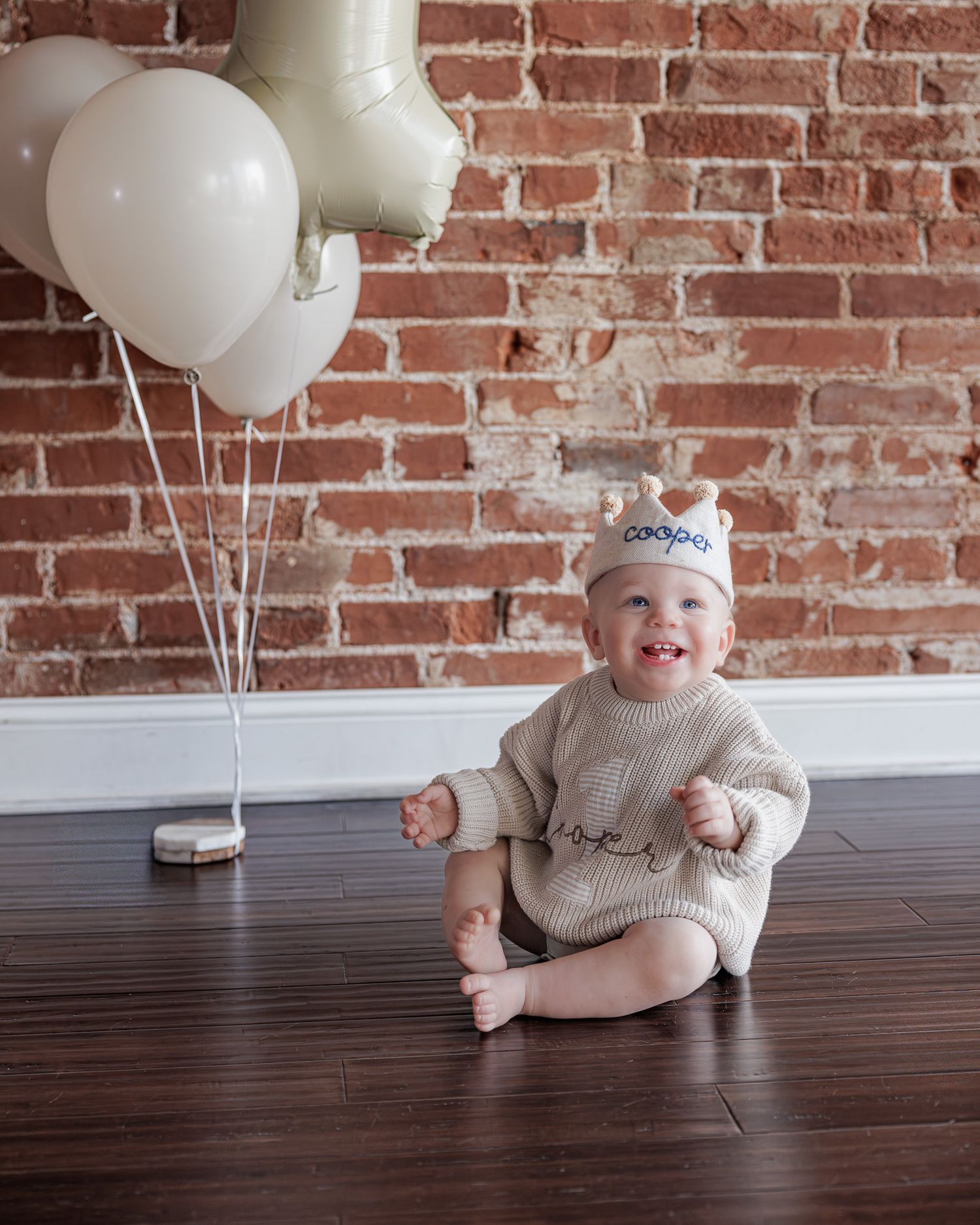 Smiling baby in beige knit romper and birthday hat sits on wood floor with white balloons against brick wall.