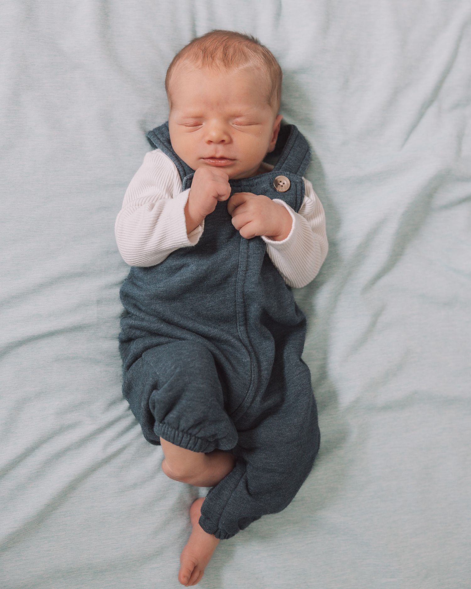 Sleeping newborn baby in blue overalls and white onesie lying on a light blue blanket.