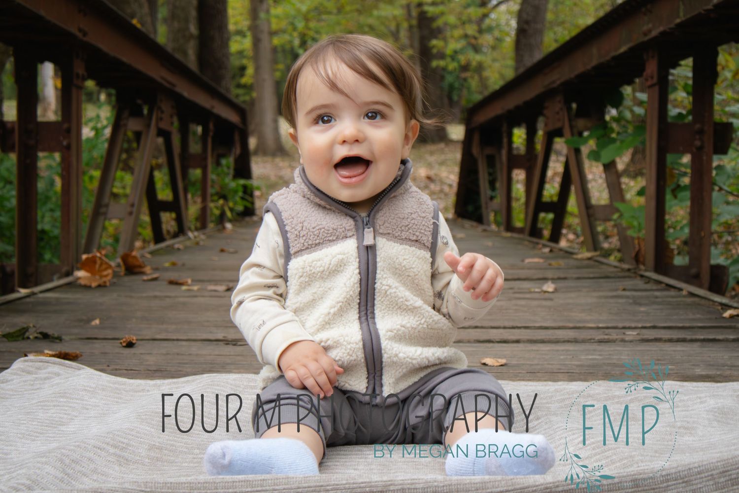 A small child wearing a gray zip-up sweater crawls and explores on a wooden bridge walkway in a forest setting.