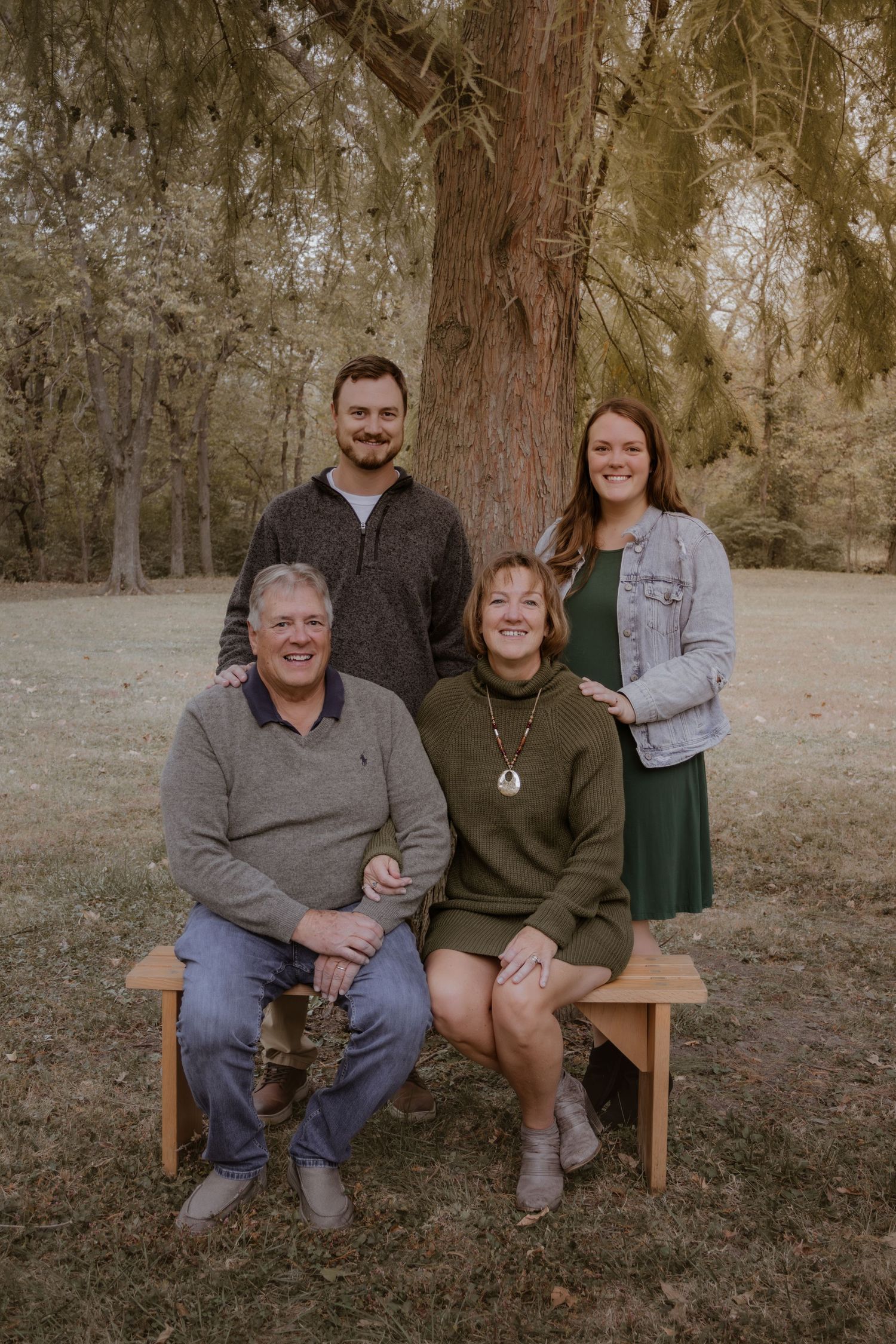 A family of four poses together outdoors on a wooden bench with autumn trees in the background.