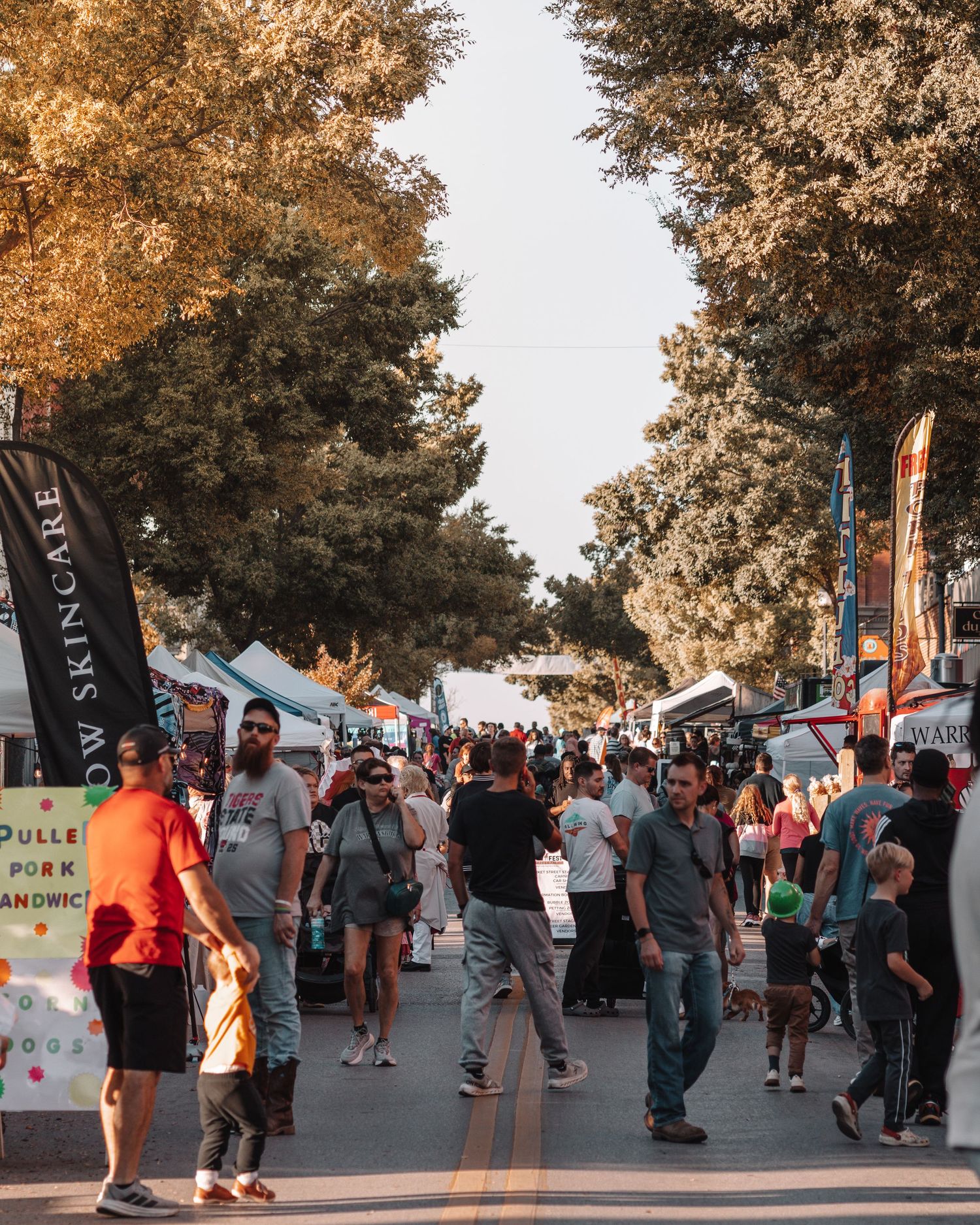 People gathering at an outdoor festival with vendor booths and autumn trees lining the walkway.
