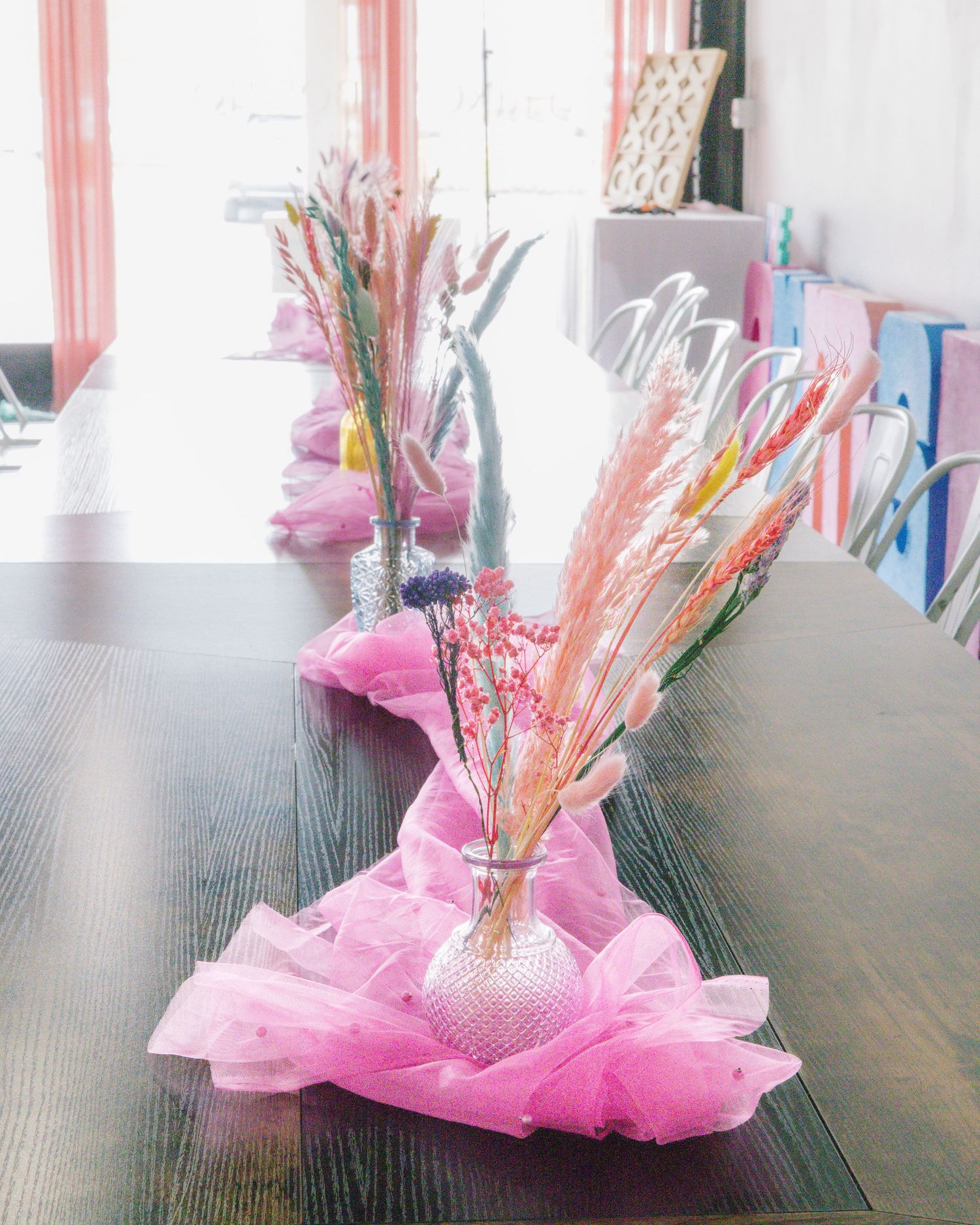 Pink tulle-wrapped glass vases with colorful dried floral arrangements as wedding table centerpieces on dark wood table.