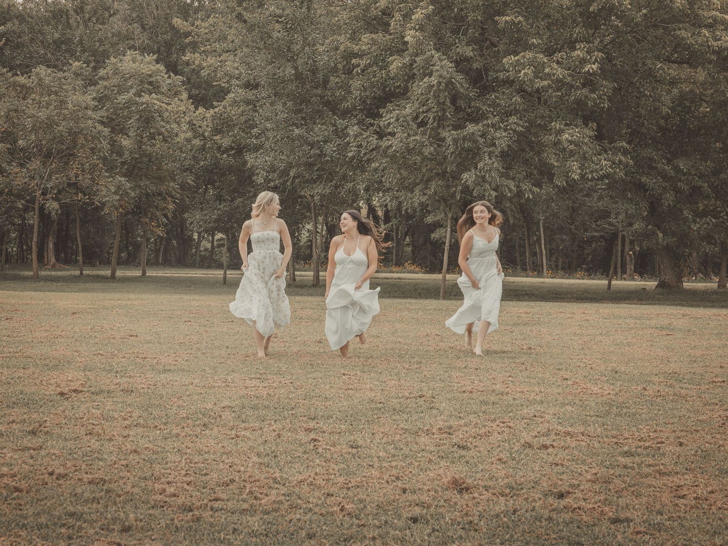 Three bridesmaids in light flowy dresses running joyfully through a lush green park on a warm sunny day.