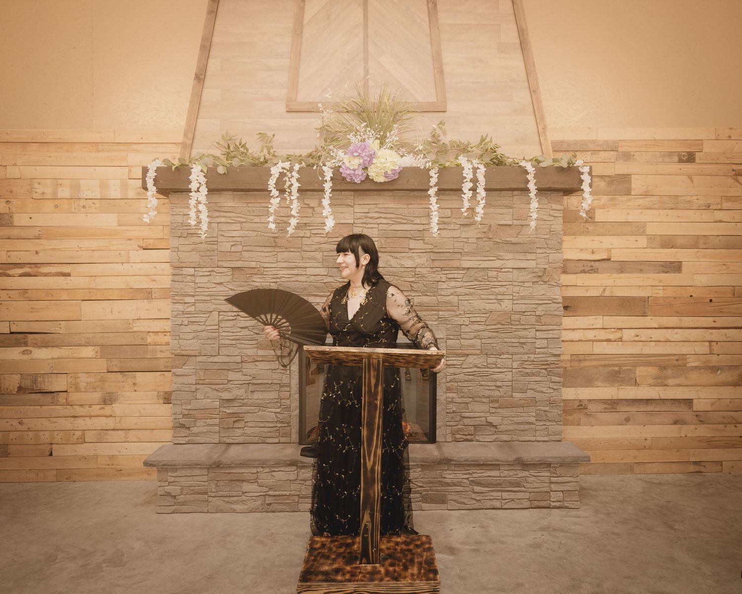 Woman in black gown holding fan speaks at ornate wooden podium before stone fireplace adorned with floral garlands.