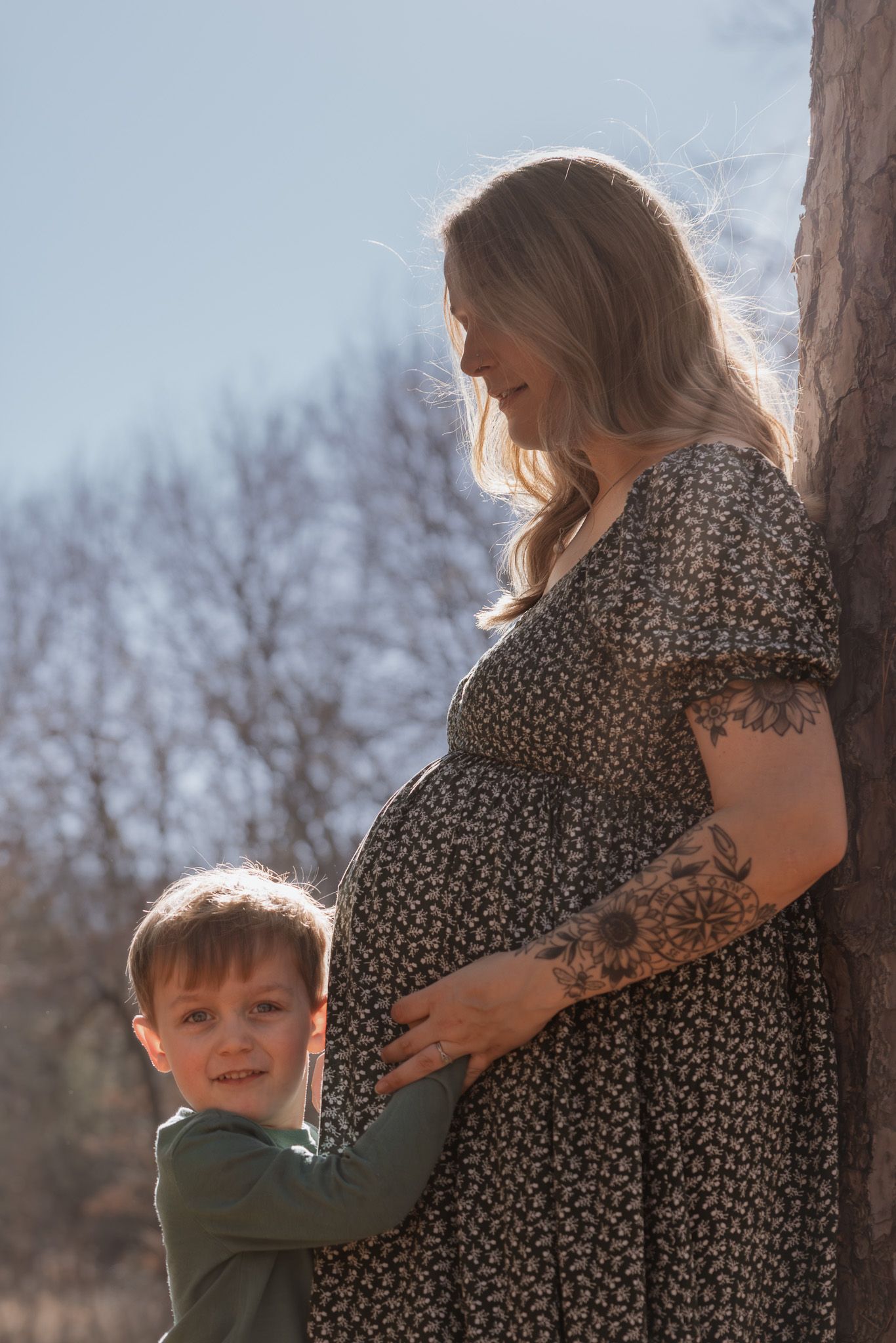 A young boy hugs his pregnant mother's belly outdoors, as she smiles down at him near a tree in soft sunlight.