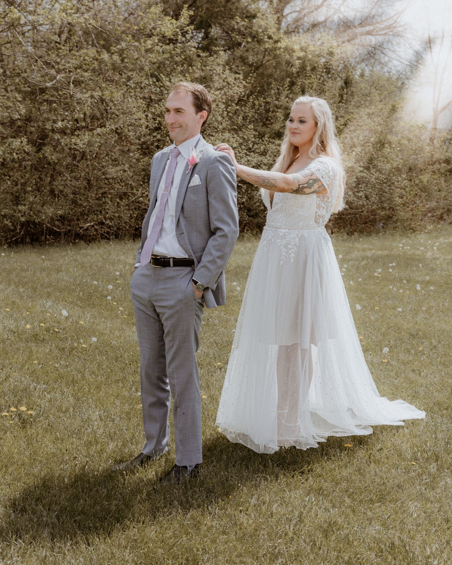 A bride in a white lace gown taps a groom in a gray suit on the shoulder during an outdoor first look.