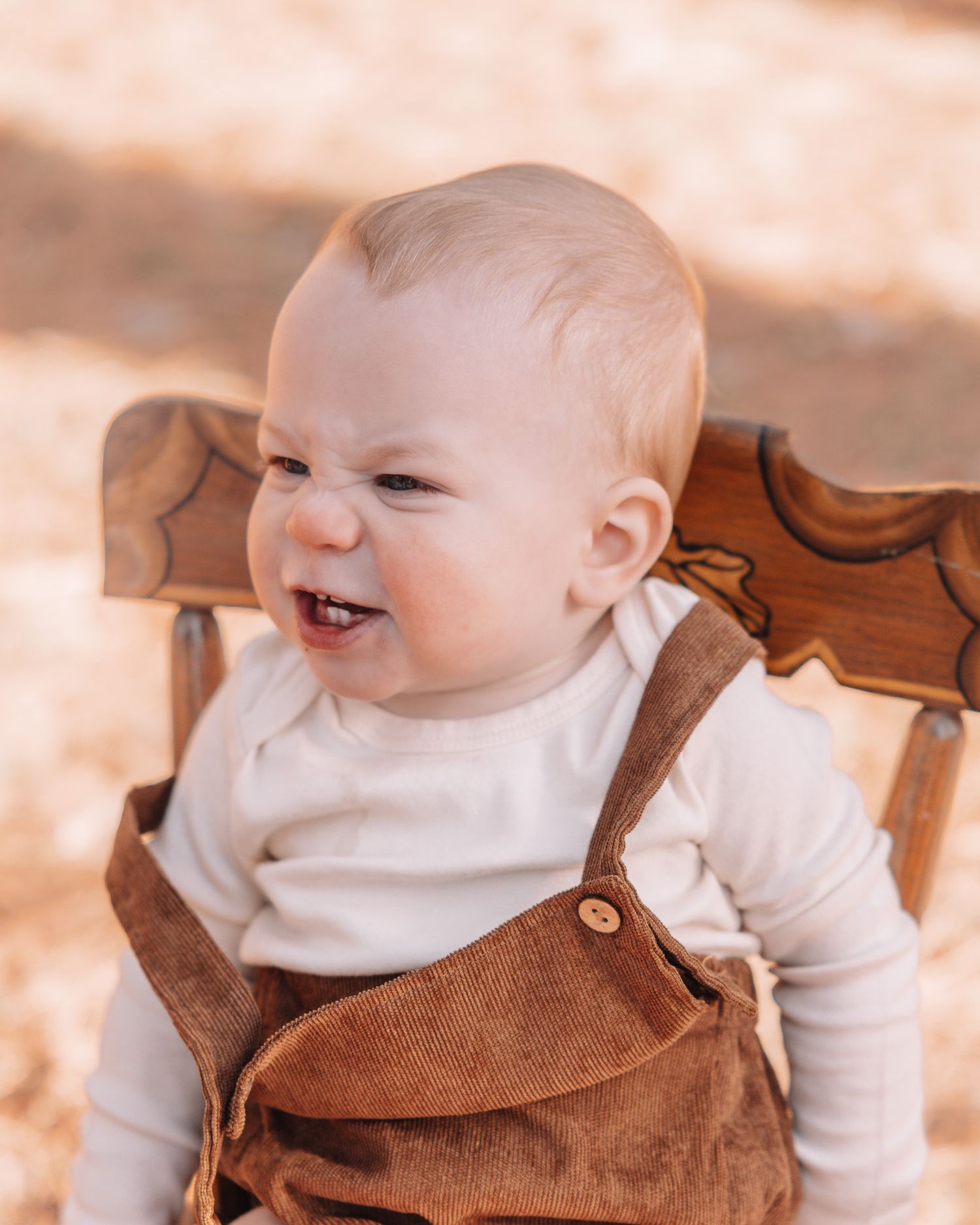 Baby in white shirt and brown overalls smiles while sitting in wooden chair.