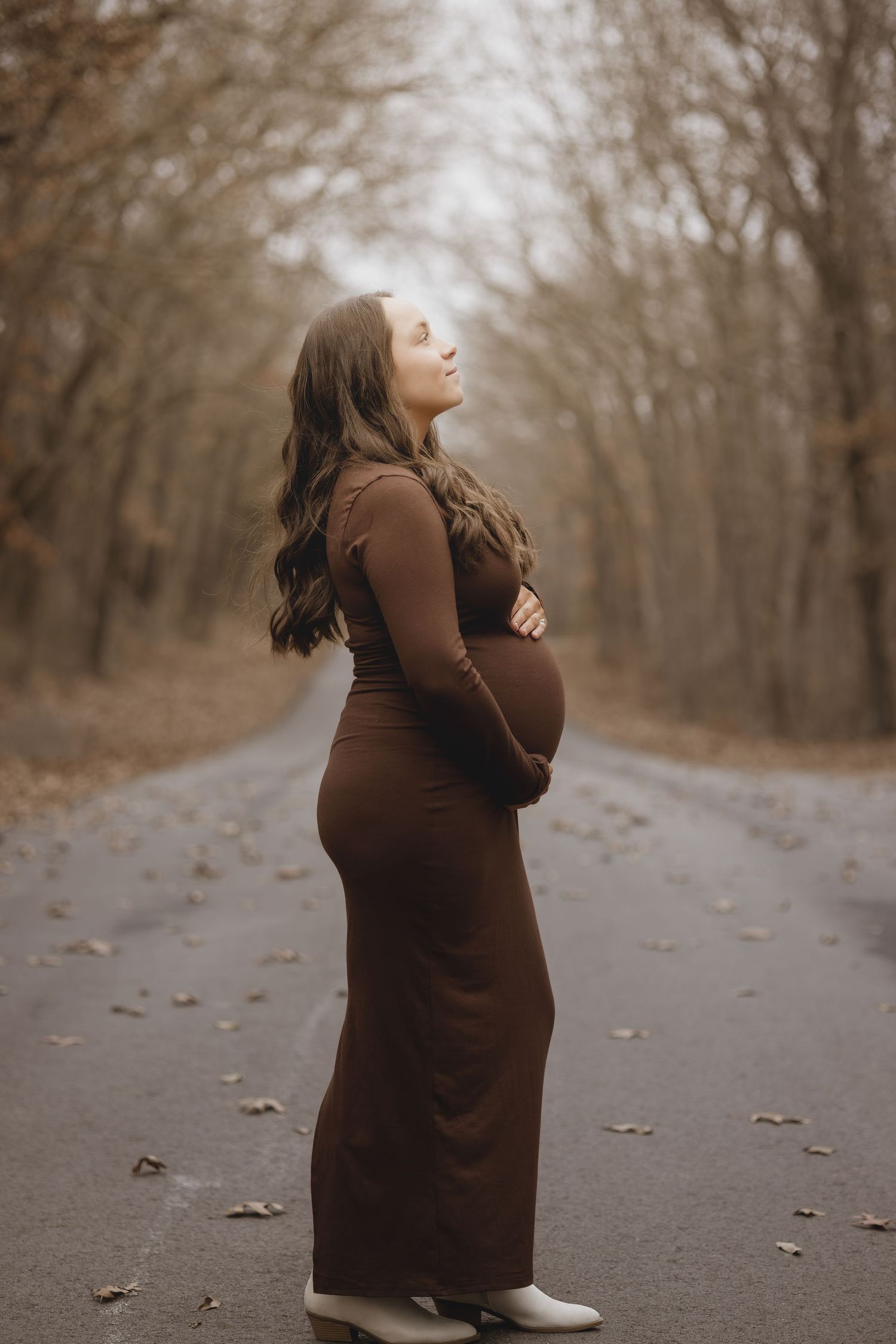 Pregnant woman in brown maxi dress standing on autumn road, cradling bump, surrounded by bare trees.