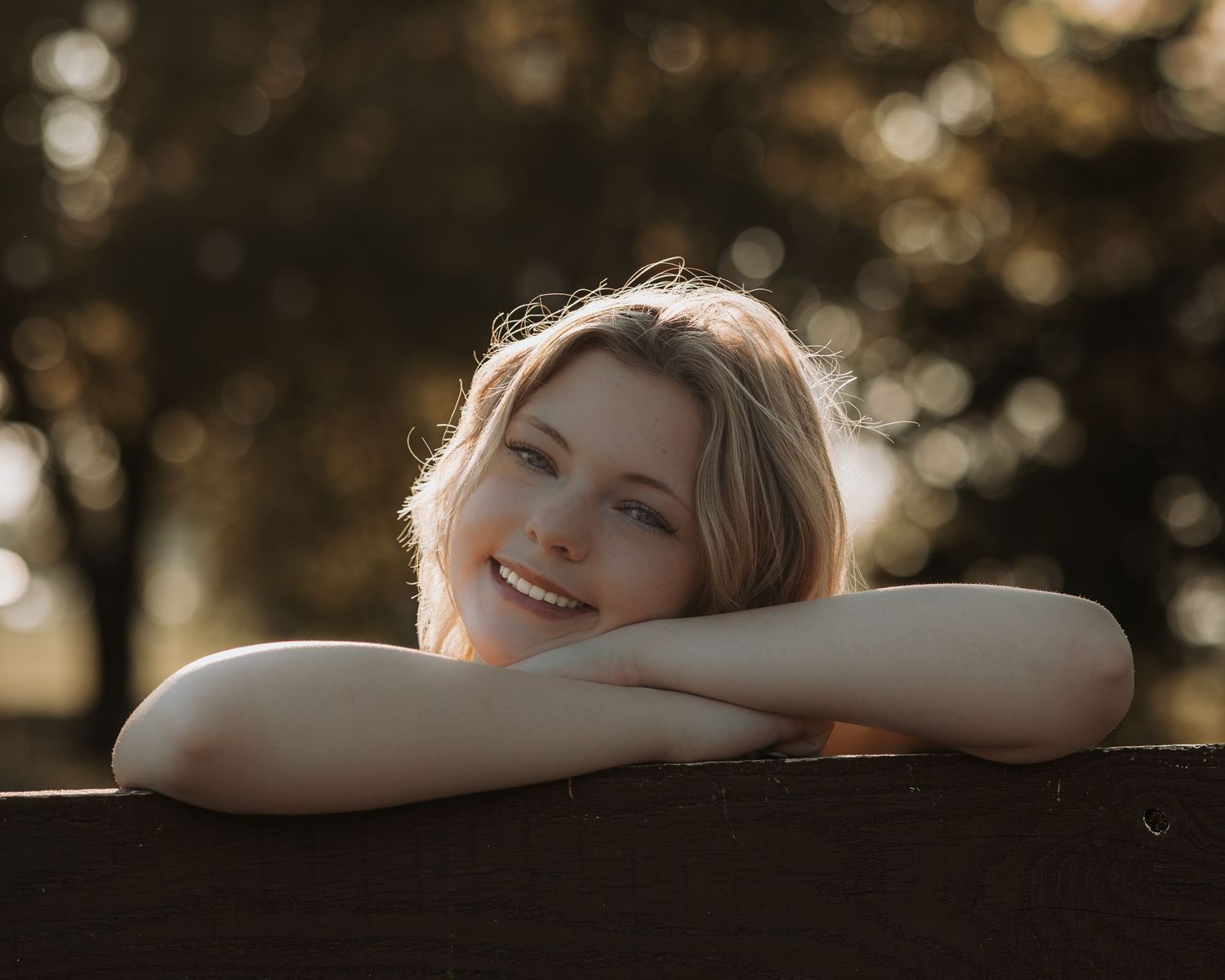 A woman rests her arms on a wooden surface outdoors during golden hour with a warm bokeh background.