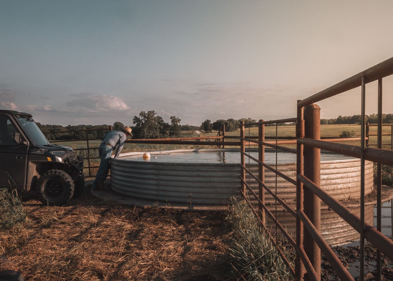 Farmer checking a large circular metal water tank on a rural farm at dusk near a gate and utility vehicle.