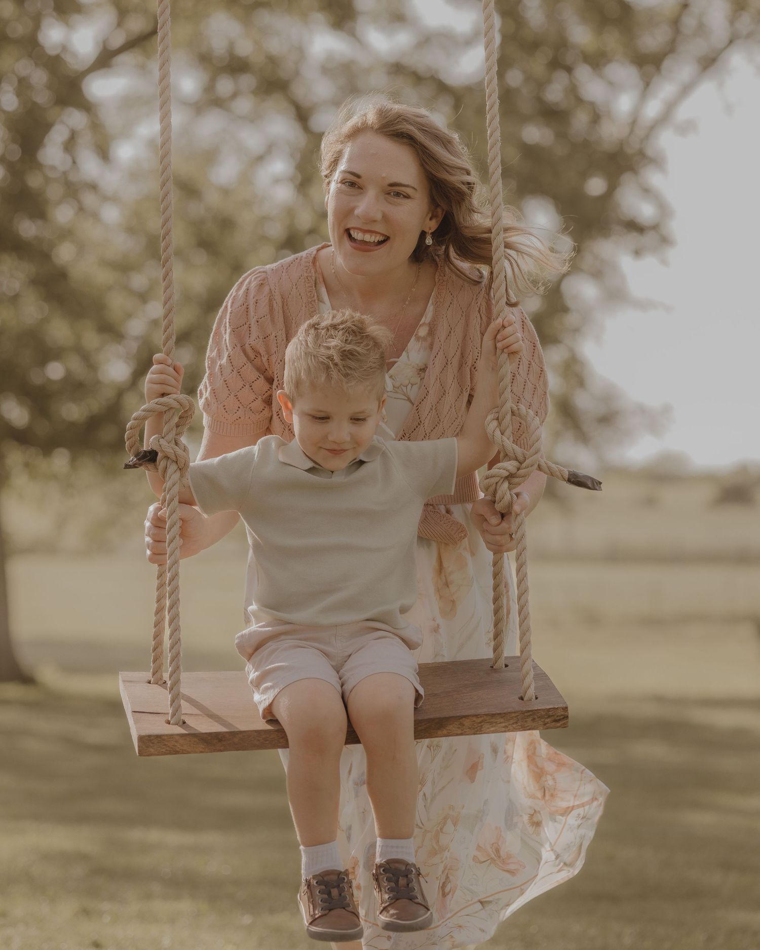 A laughing mother in a pink cardigan pushes her young son on a rustic wooden rope swing outside.