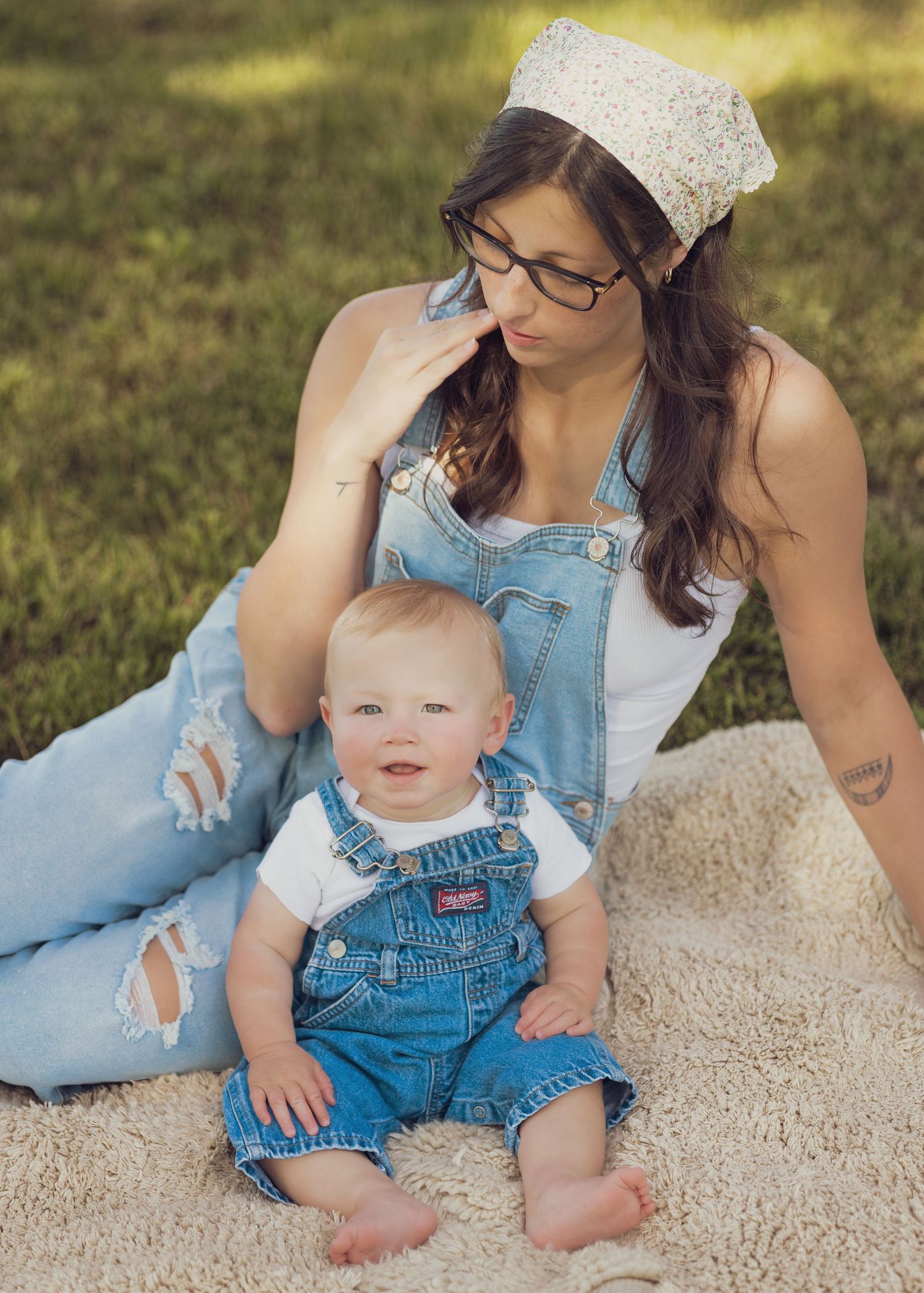 A young mother in denim overalls sits on a blanket outdoors with her smiling baby boy also wearing overalls.